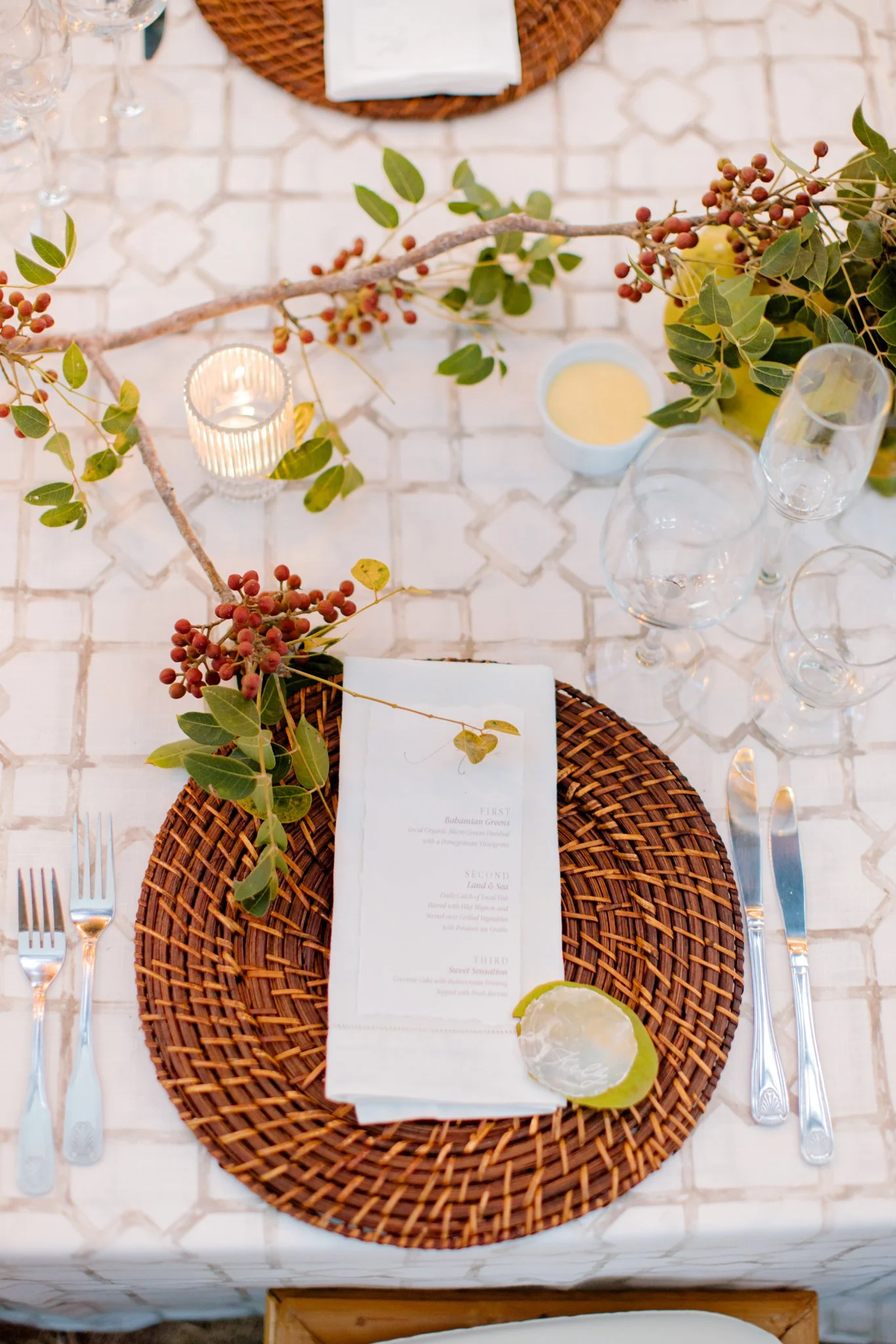 A formal table setting with a wicker charger, a white napkin, a menu, a lemon wedge, and greenery with berries. The table has glassware, a lit candle, and a small bowl of sauce or dressing, all on a white tiled table surface.