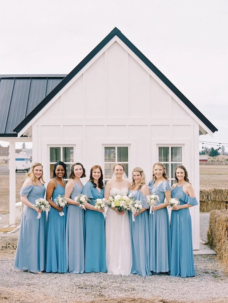 Bride in a white wedding dress and seven bridesmaids in matching blue dresses standing in front of a small white house with a black roof, each holding a bouquet of white flowers.