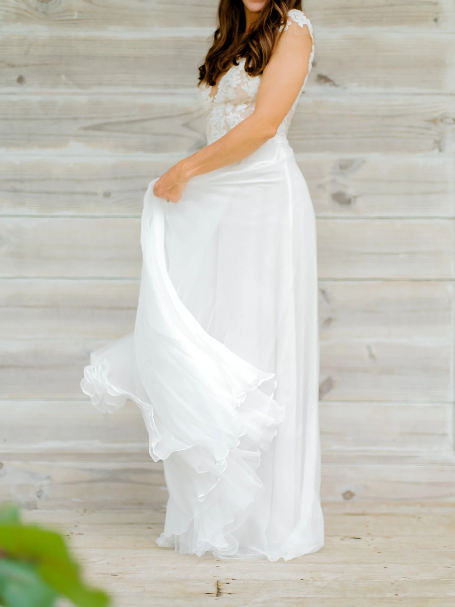 A woman in a white lace and chiffon dress standing against a light wooden wall.