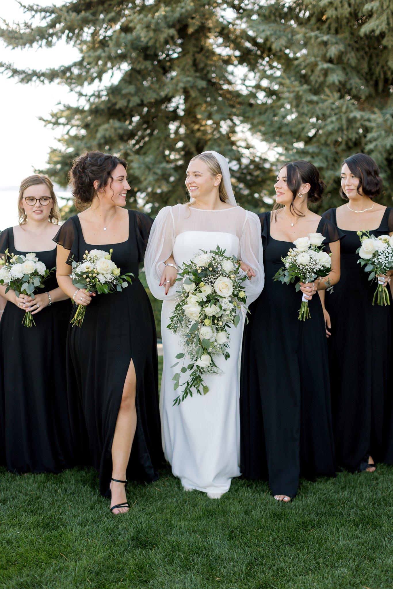 Bride walking with her bridesmaids holding white floral bouquets on the lawn during a summer wedding in Coeur d’Alene.