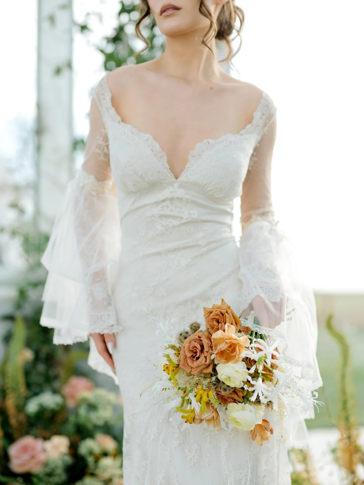 A woman in a white lace wedding gown holding a bouquet of peach, cream, and yellow flowers with greenery in a bright, outdoor setting at Greenhouse on Harvard near Spokane, Washington