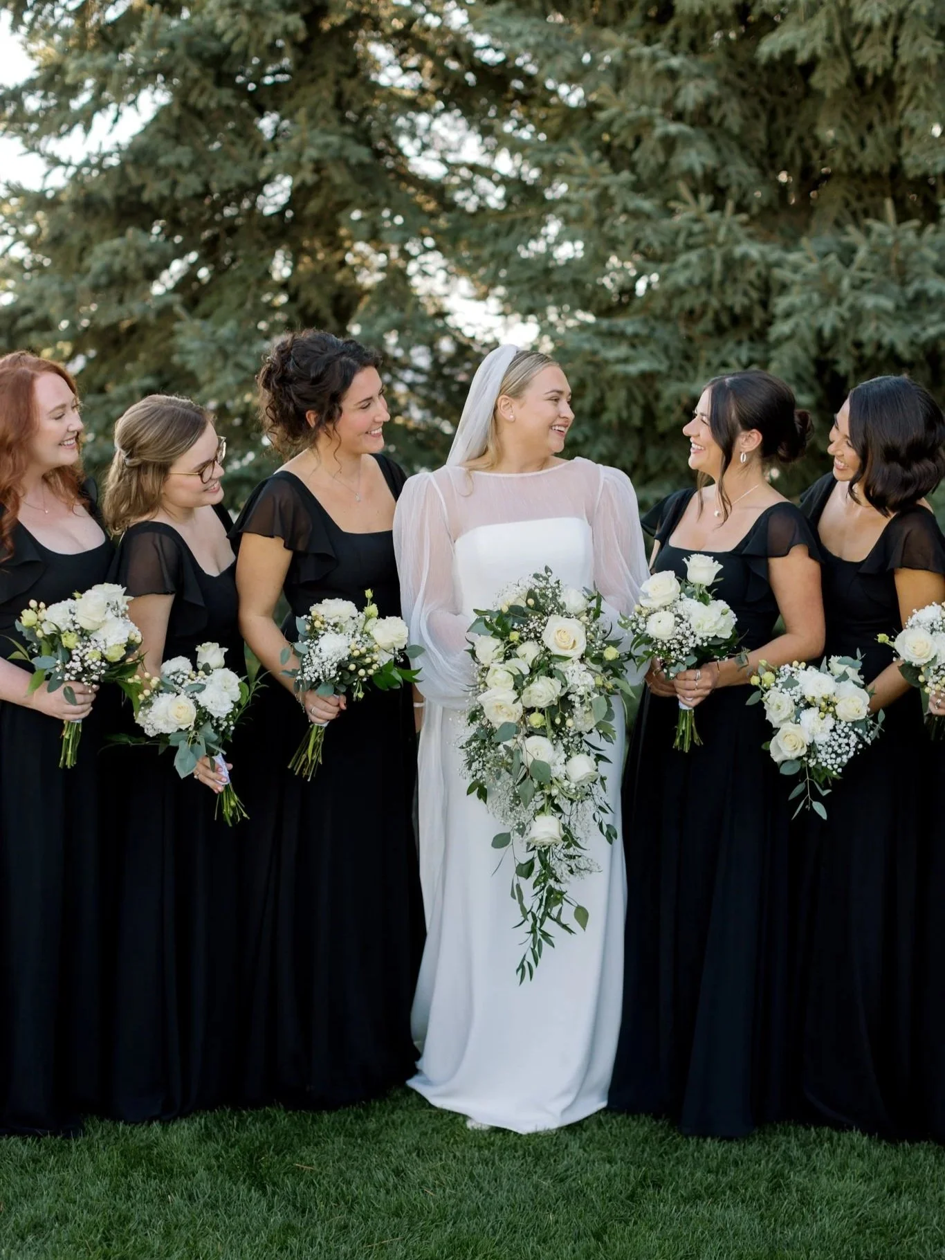 Bride smiling with her bridesmaids in black dresses holding white bouquets in front of evergreen trees at a Hagadone Event Center wedding.