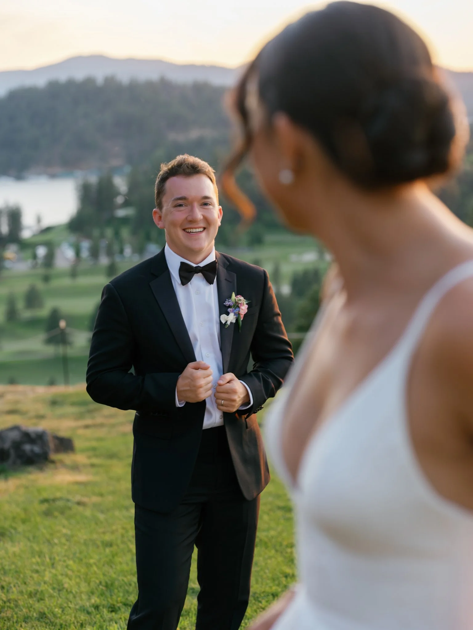 A groom in a tuxedo smiling and looking at a bride who is out of focus, against a scenic outdoor backdrop with mountains and a lake during sunset at Elk Point wedding above Lake Coeur d’Alene.