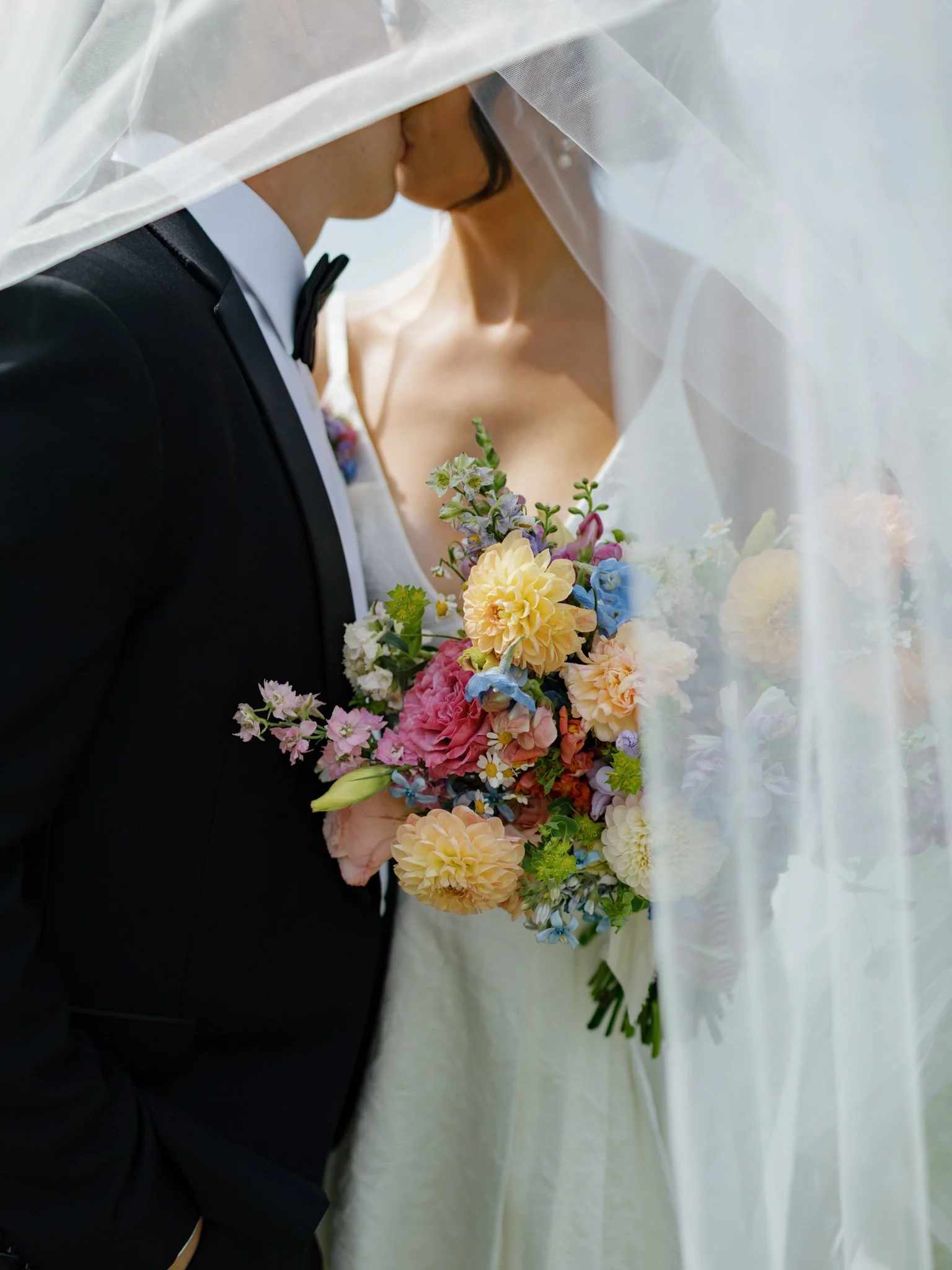 A bride and groom kissing under a wedding veil, with the bride holding a colorful bouquet of flowers at Elk Point wedding above Lake Coeur d’Alene.