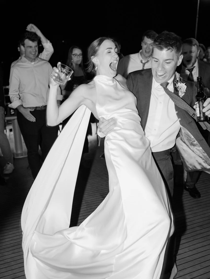 Black and white photo of a wedding celebration with a bride and groom dancing closely, surrounded by smiling guests.