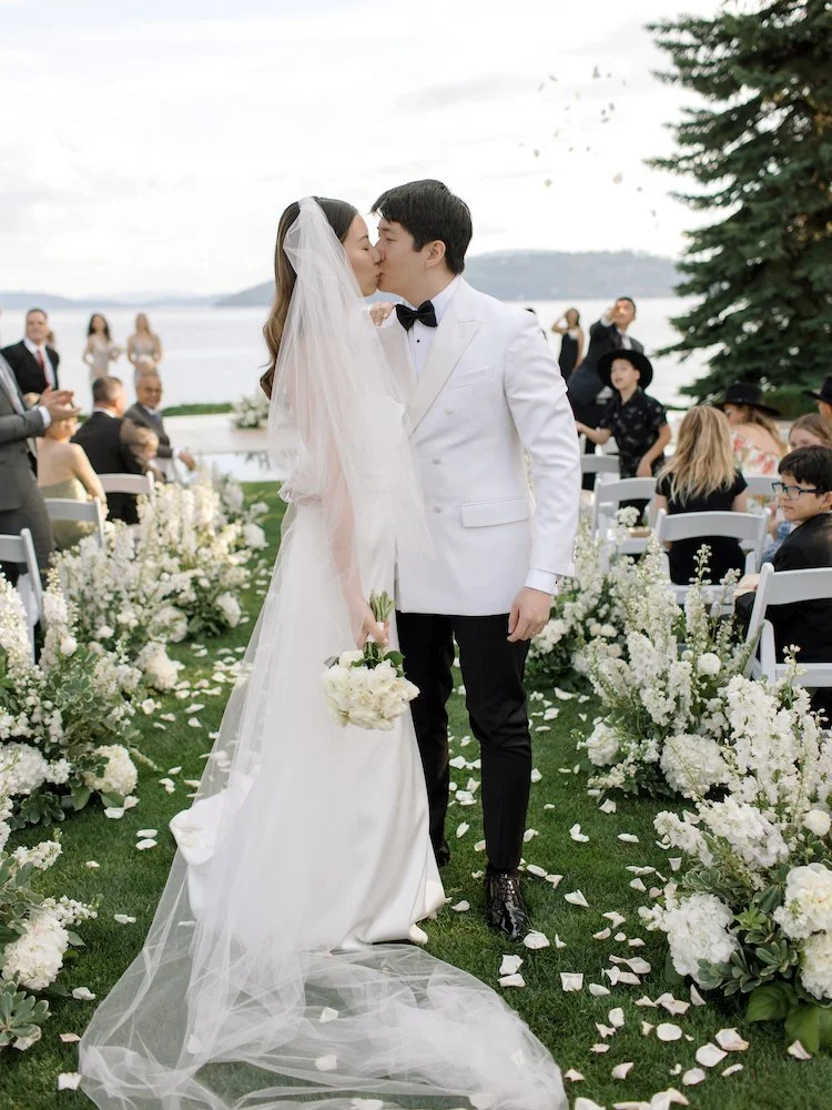 Bride and groom kiss during outdoor wedding ceremony with guests and floral decorations.
