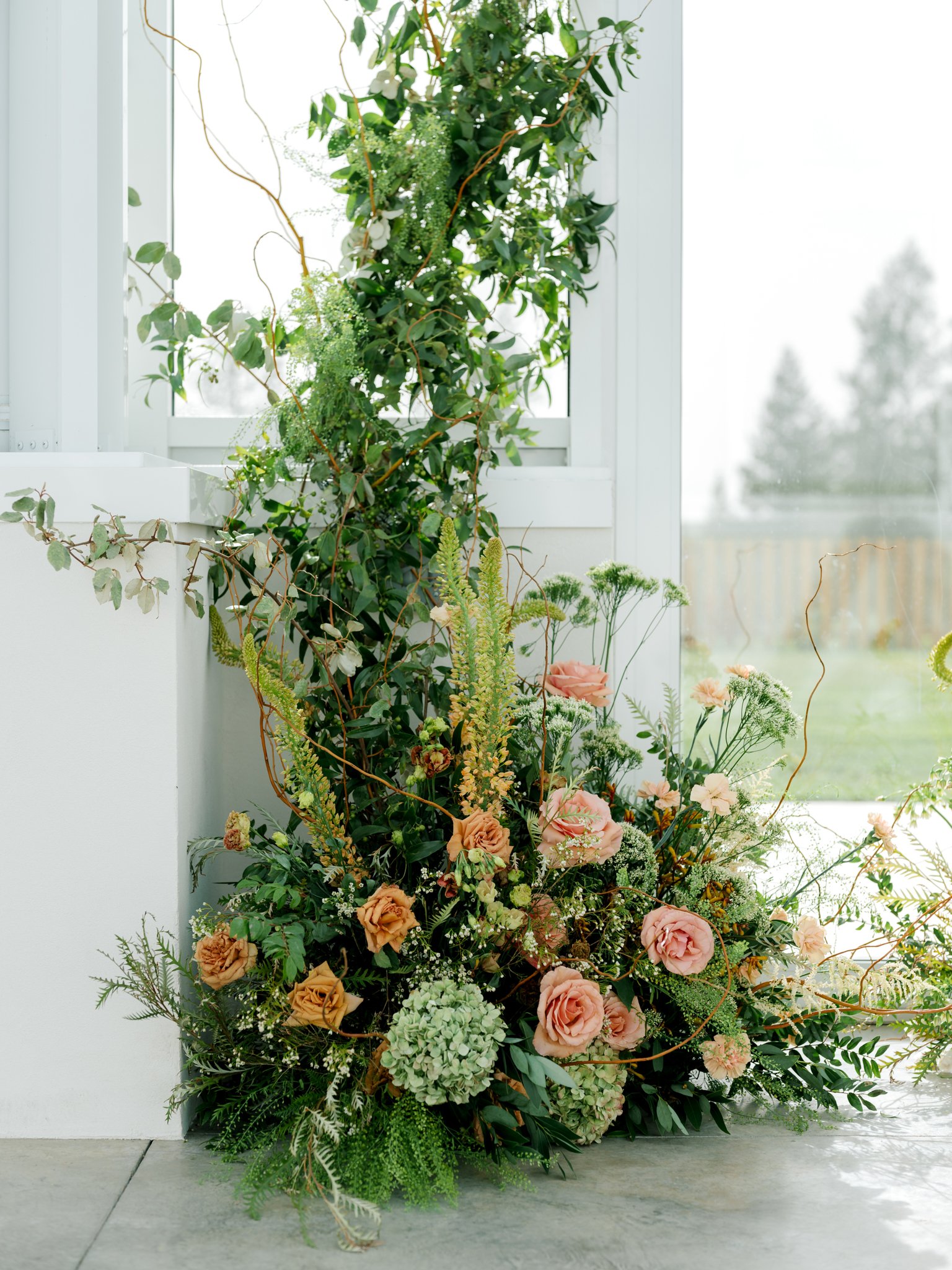 A floral arrangement with peach roses, white hydrangeas, and various greenery near a window with a view of a backyard at Greenhouse on Harvard near Spokane, Washington.