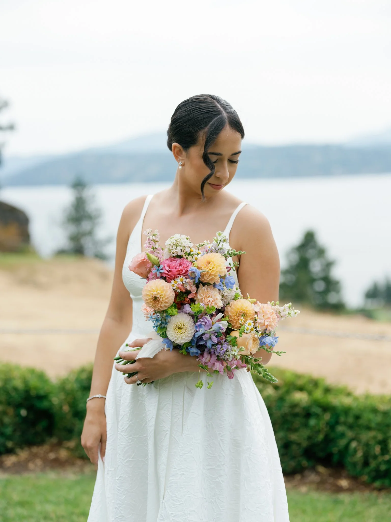 A bride in a white dress holding a colorful bouquet of flowers with a lake and mountains in the background at Elk Point wedding in Coeur d’Alene.