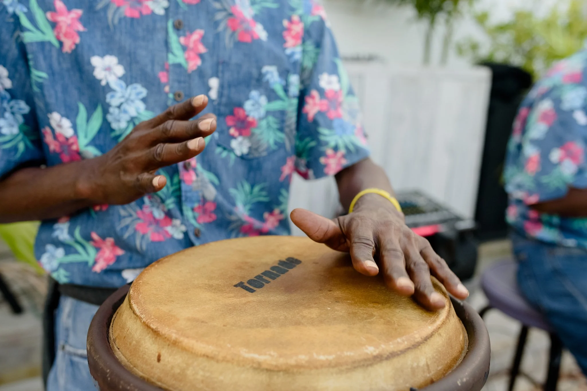Person playing conga drum wearing a blue floral shirt, with another person in similar attire in the background.