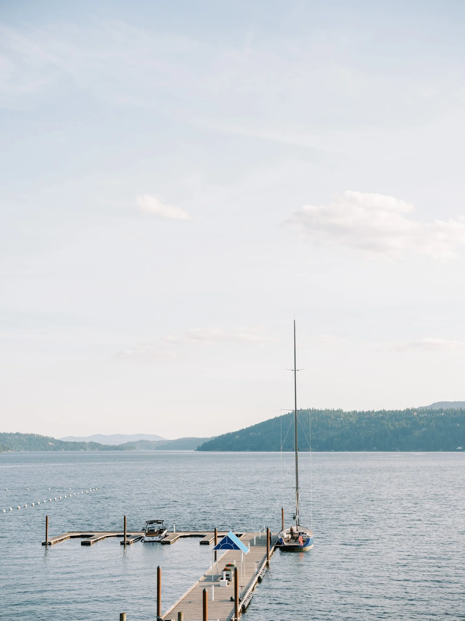 A dock extending into a calm lake with boats moored, surrounded by hills under a partly cloudy sky at the Hagadone Event Center in Coeur d'Alene, Idaho.