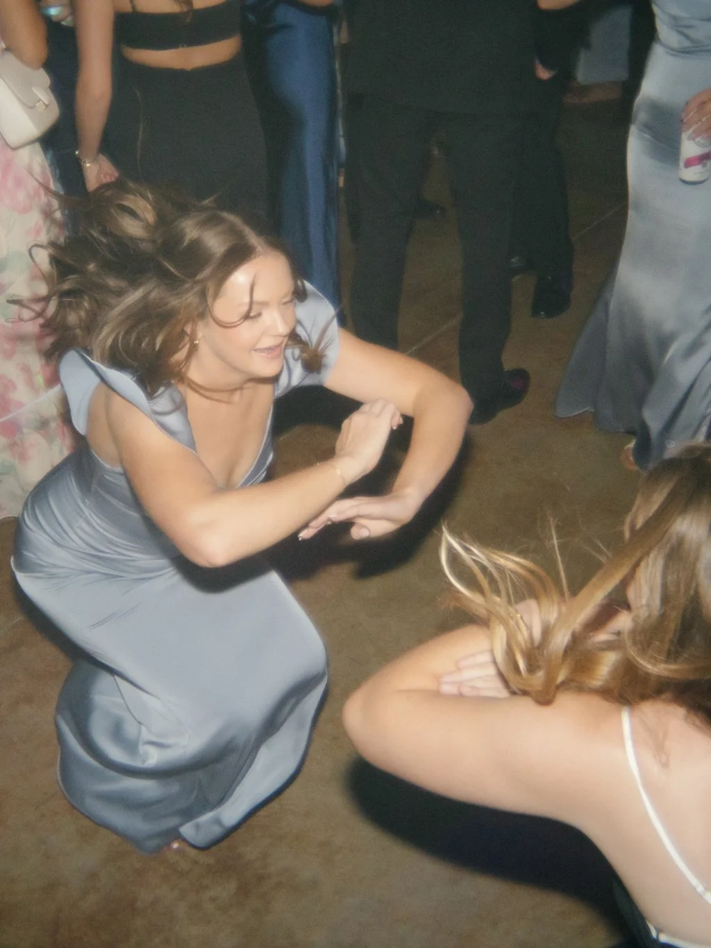 A woman in a silver dress is dancing at a party with others, while others stand around in the background.