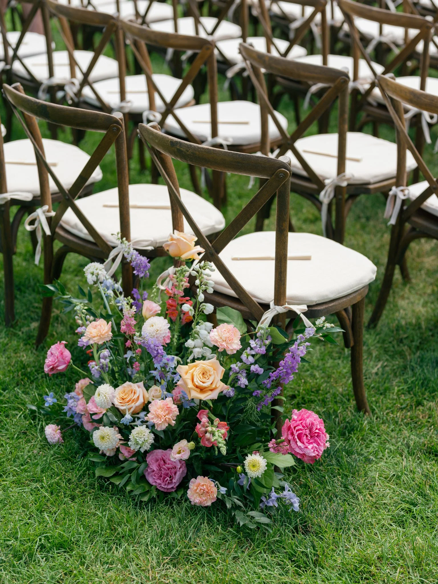 A floral arrangement with pink and peach roses, purple and white flowers, and green foliage placed on grass, with wooden chairs arranged in rows in the background, likely for an outdoor wedding at Elk Point wedding in Coeur d’Alene.