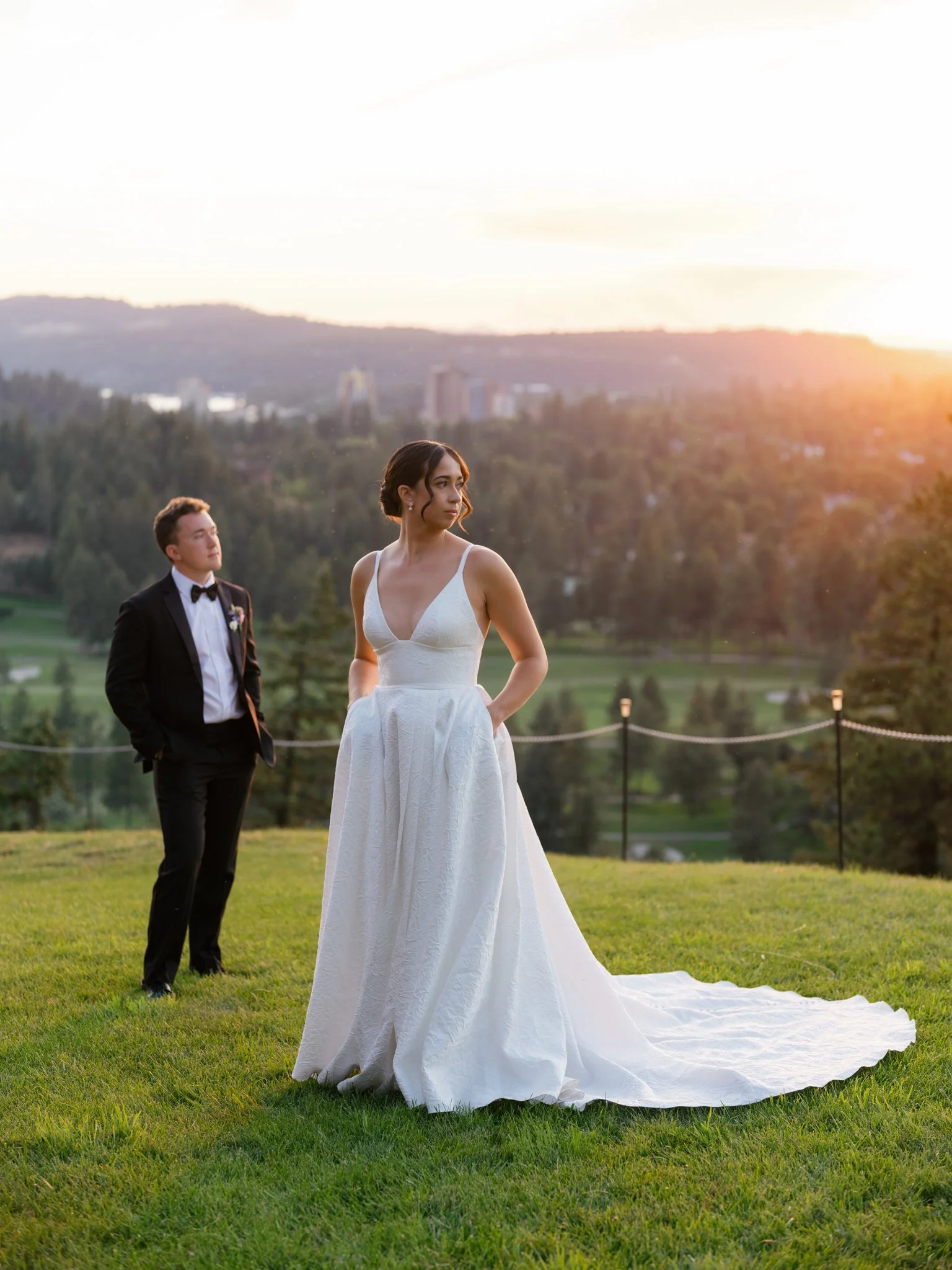 Bride in a white wedding dress stands on a grassy lawn during sunset, with a groom in a black tuxedo standing behind her, in a scenic outdoor setting at Elk Point wedding above Lake Coeur d’Alene.