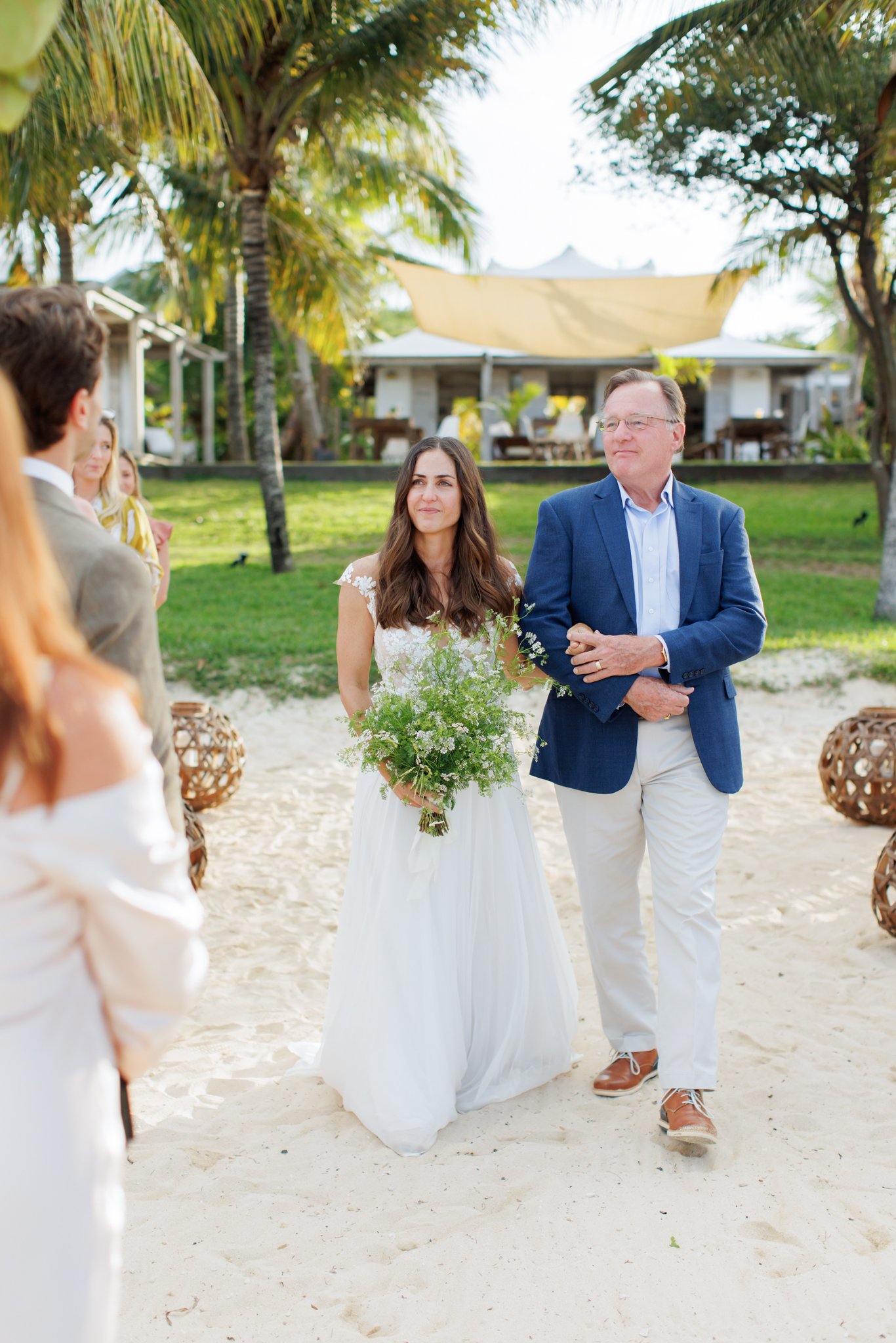 A bride with long dark hair wearing a white wedding dress holding a bouquet of flowers walks down the aisle, accompanied by a man in a blue blazer and white pants during a beach wedding ceremony, with guests in the background and palm trees around.