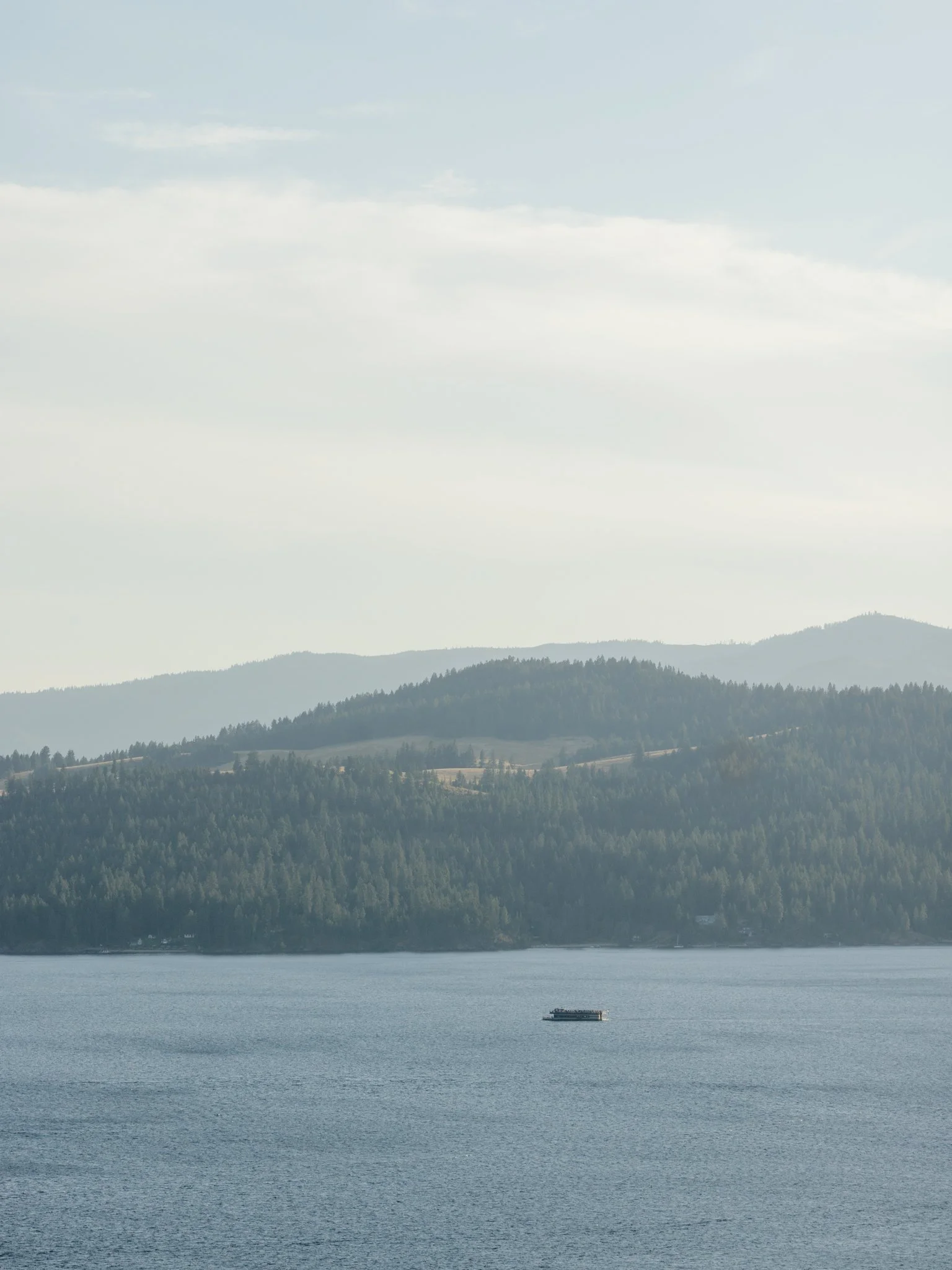 A large body of water with a boat or barge floating on it, and a background of forested hills and mountains under a partly cloudy sky at Elk Point mountaintop wedding.