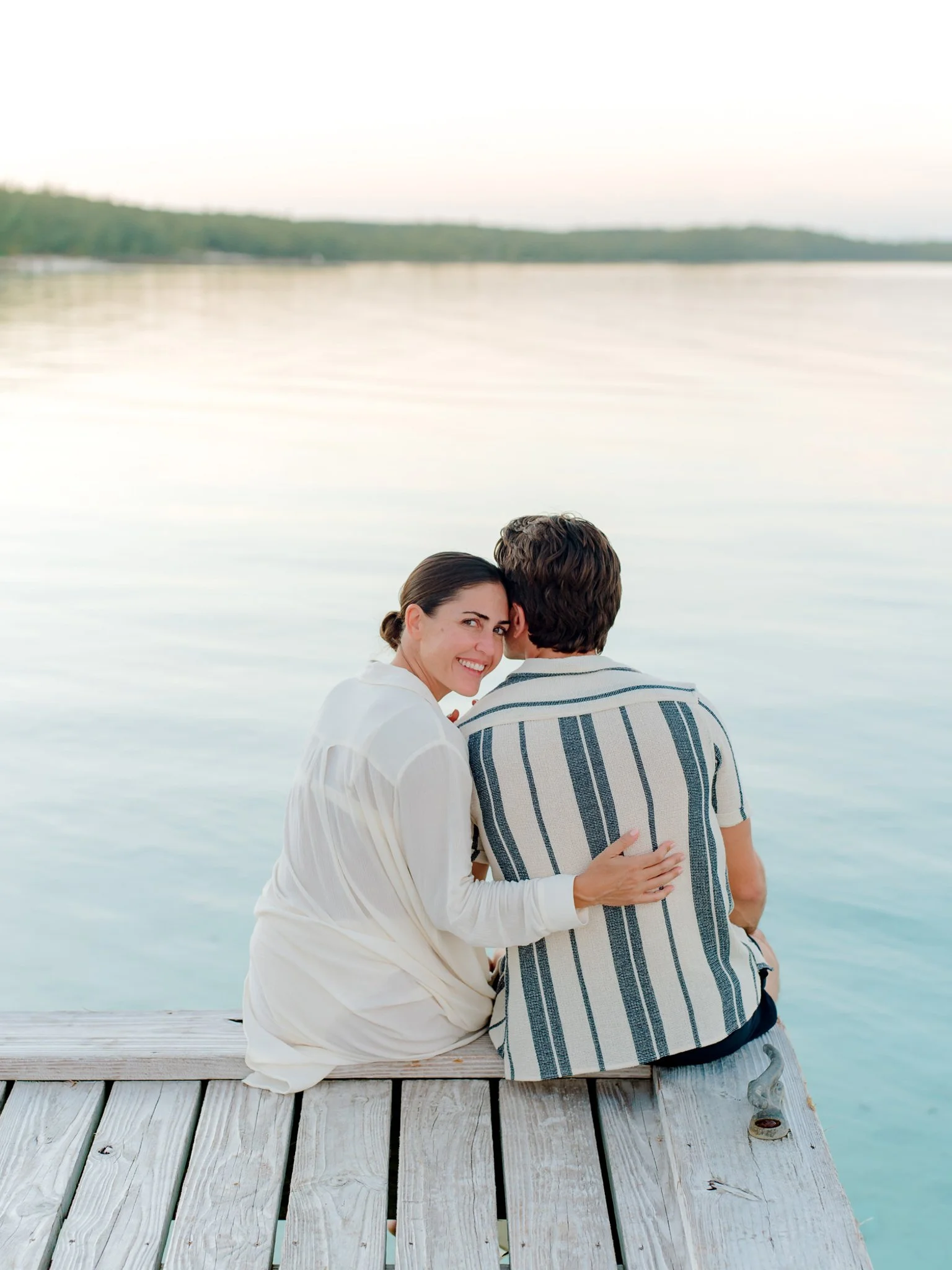 A woman smiles as she sits on a wooden dock beside a man, with their backs to the camera, by a calm body of water during sunset or sunrise at The Other Side Hotel Eleuthera during a luxury Bahamas destination wedding.