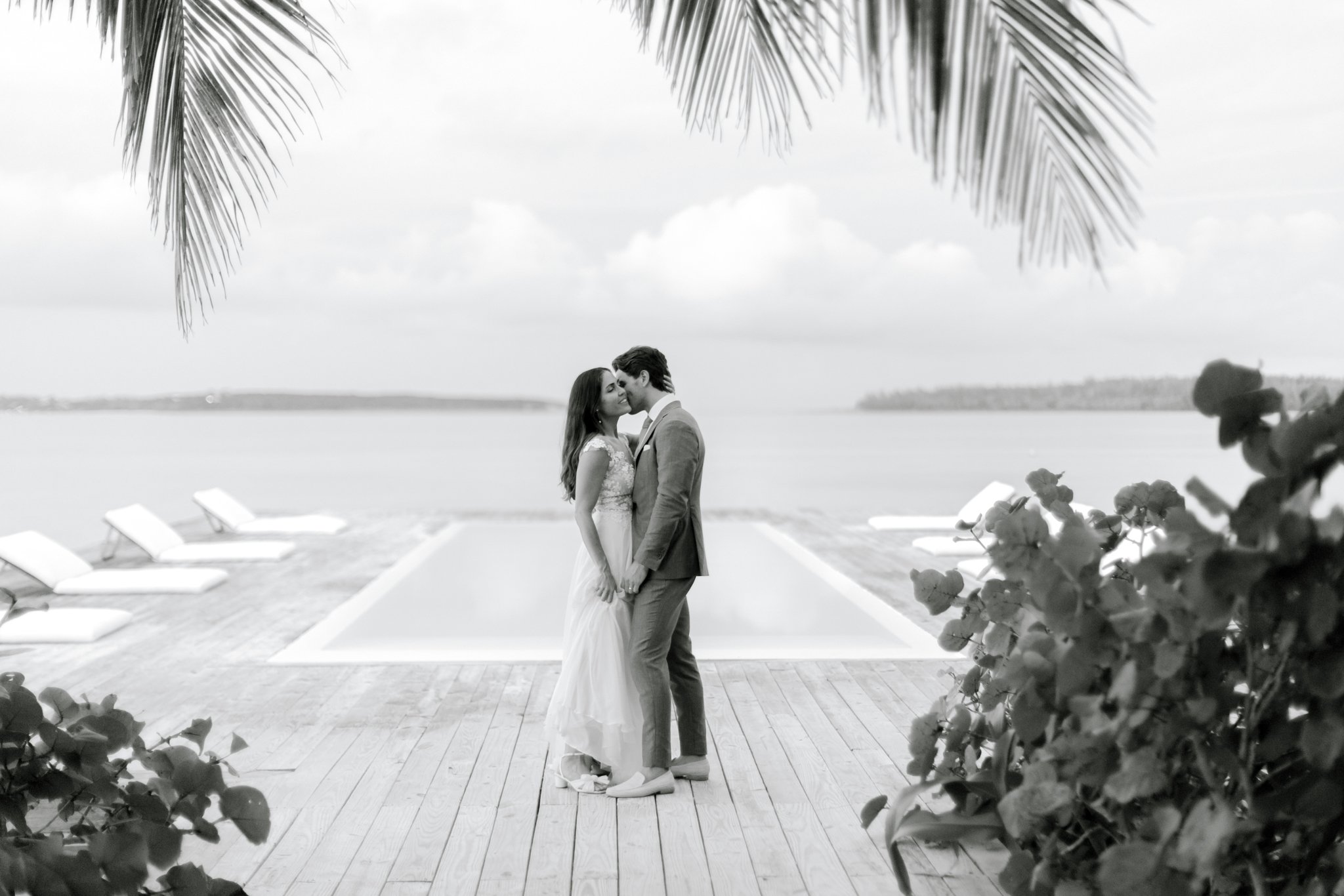 A black and white photo of a couple in wedding attire standing close on a wooden deck near water, with palm tree leaves at the top and flowers in the foreground. The couple is kissing, with a pool and lounge chairs visible behind them.