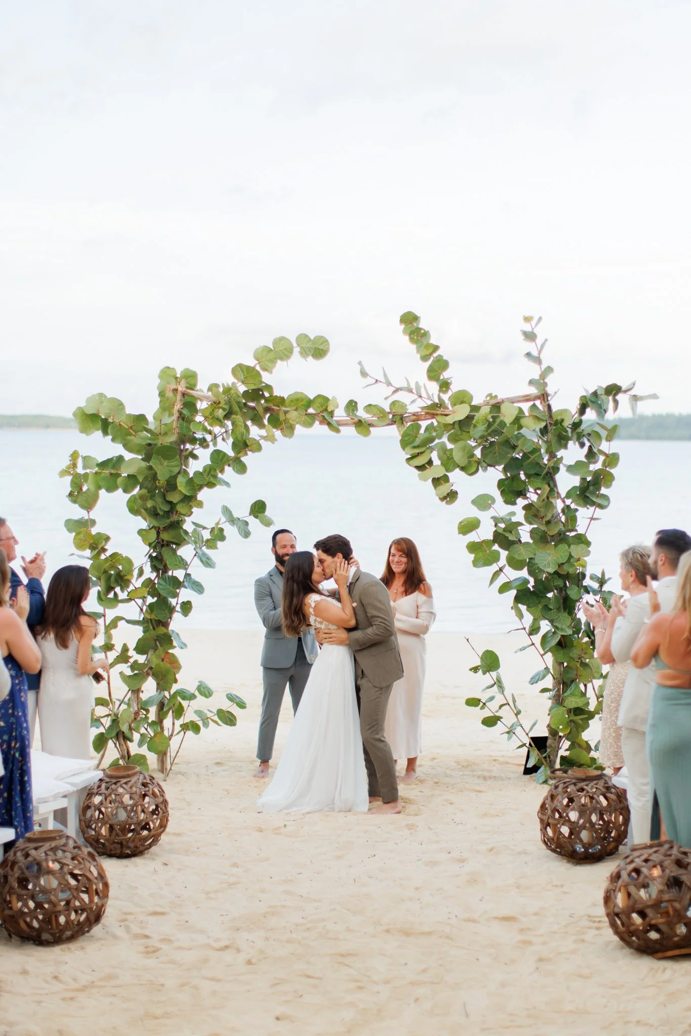 A couple getting married on a beach, kissing under a green leafy arch, with guests clapping and smiling on either side at The Other Side Hotel Eleuthera during a luxury Bahamas destination wedding.