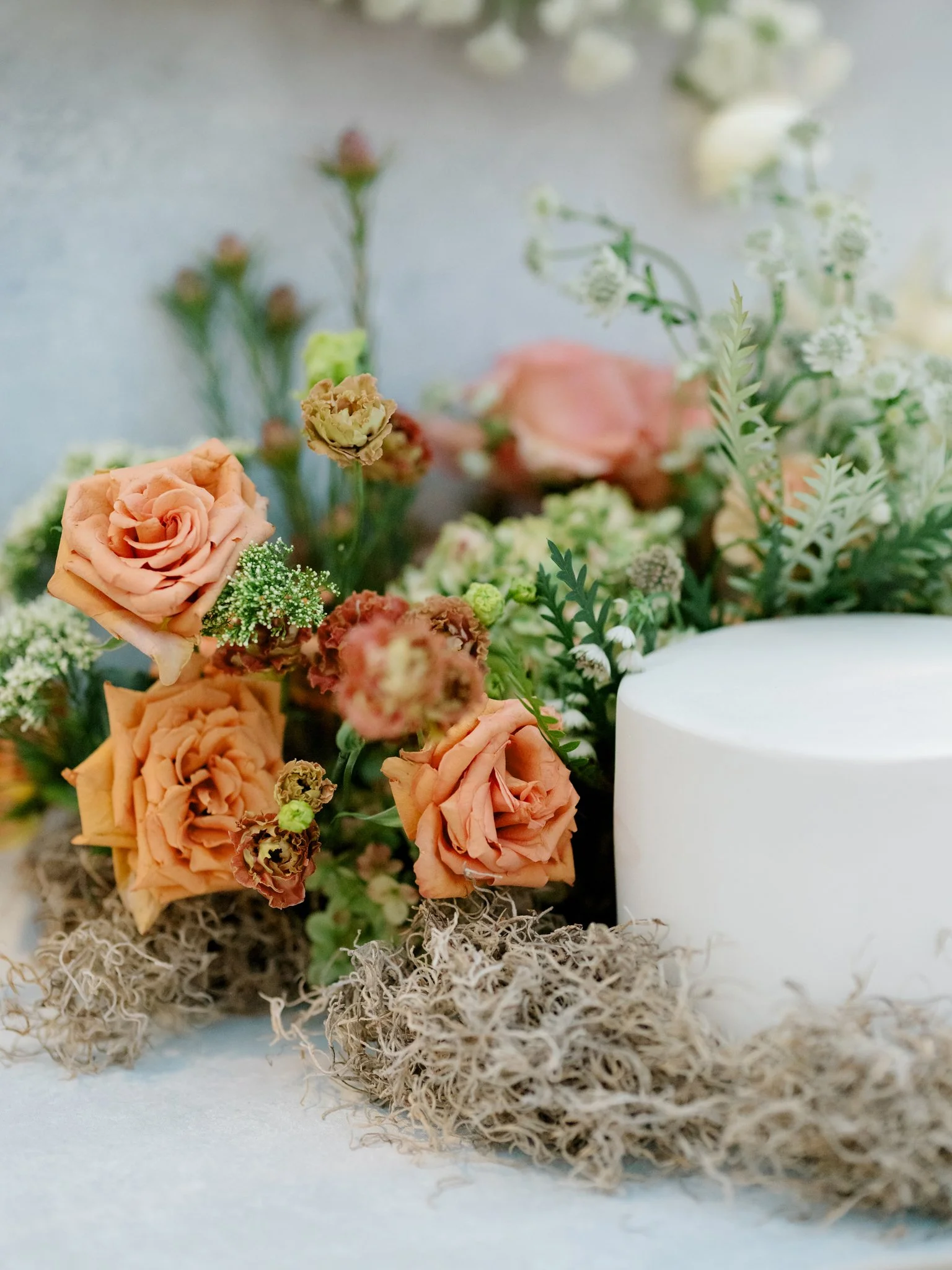 Close-up of a floral centerpiece with orange roses, pink flowers, and greenery surrounding a white candle, placed on a moss base.