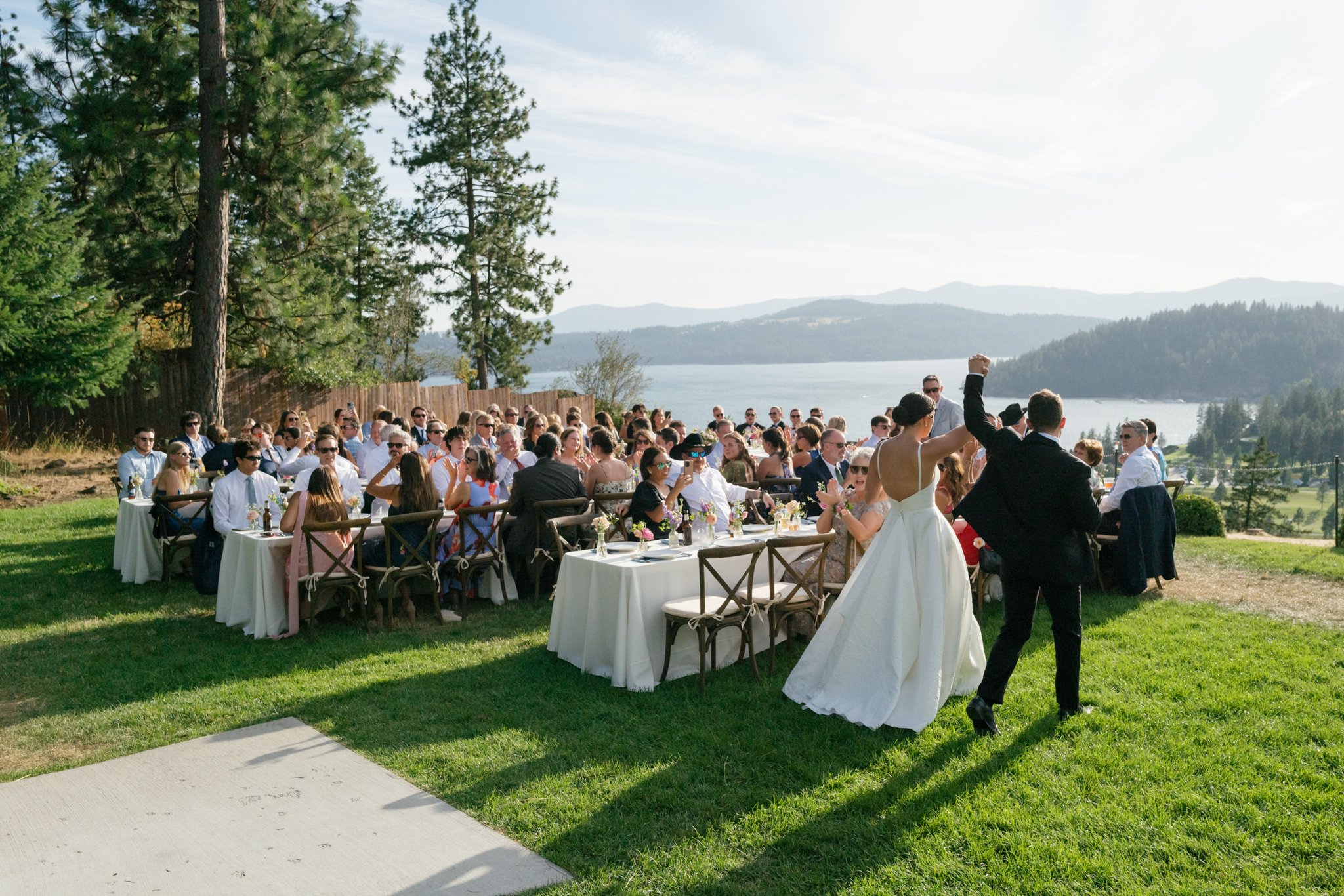A wedding reception outdoors by a lake with a large group of people sitting at long tables, a bride and groom dancing, scenic mountains and lake in the background, during daytime at Elk Point wedding above Lake Coeur d’Alene.