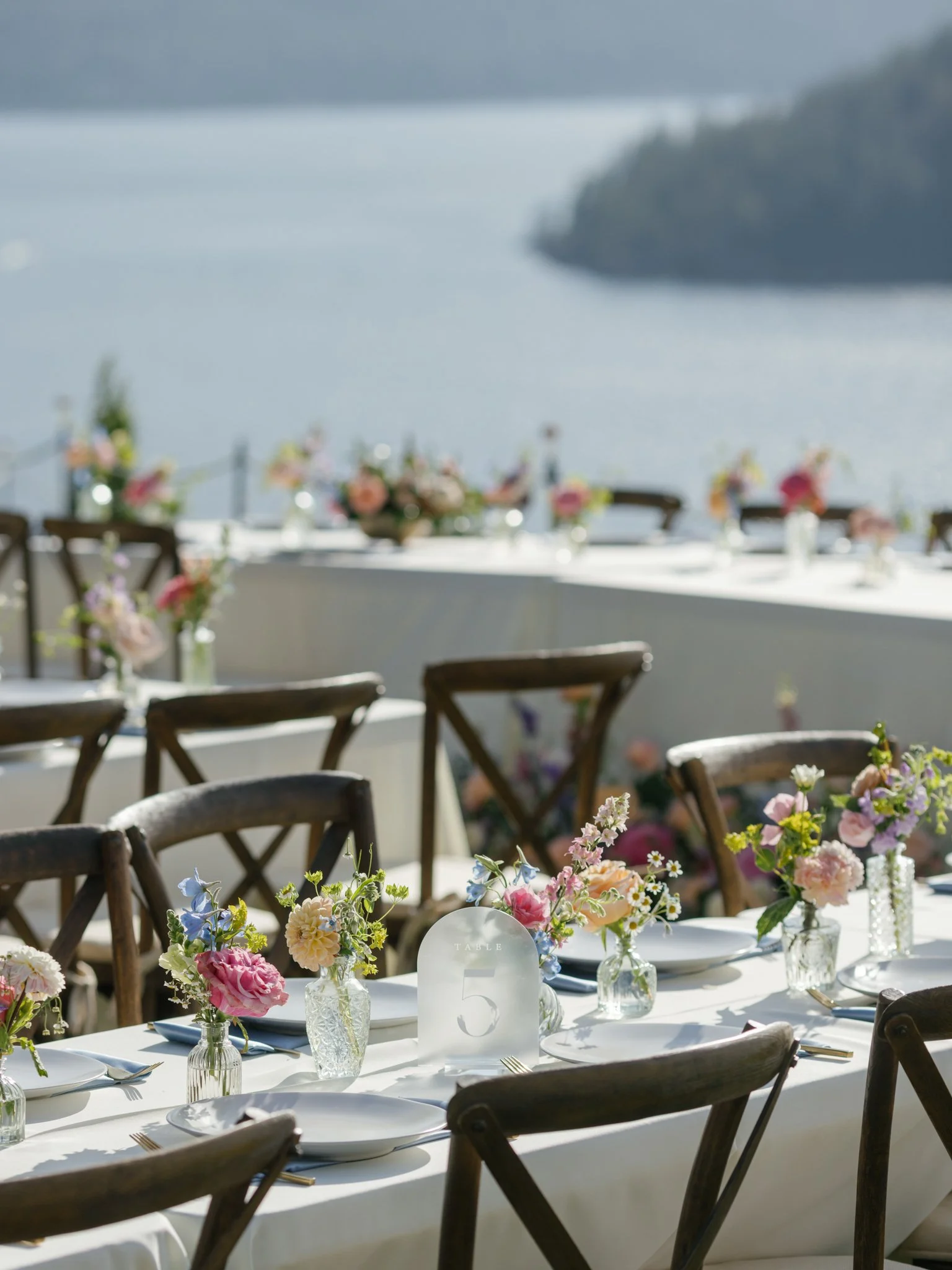 A beautifully set outdoor table decorated with pastel flower arrangements and glass vases, overlooking a body of water with mountains in the background for a special event or celebration at Elk Point wedding above Lake Coeur d’Alene.