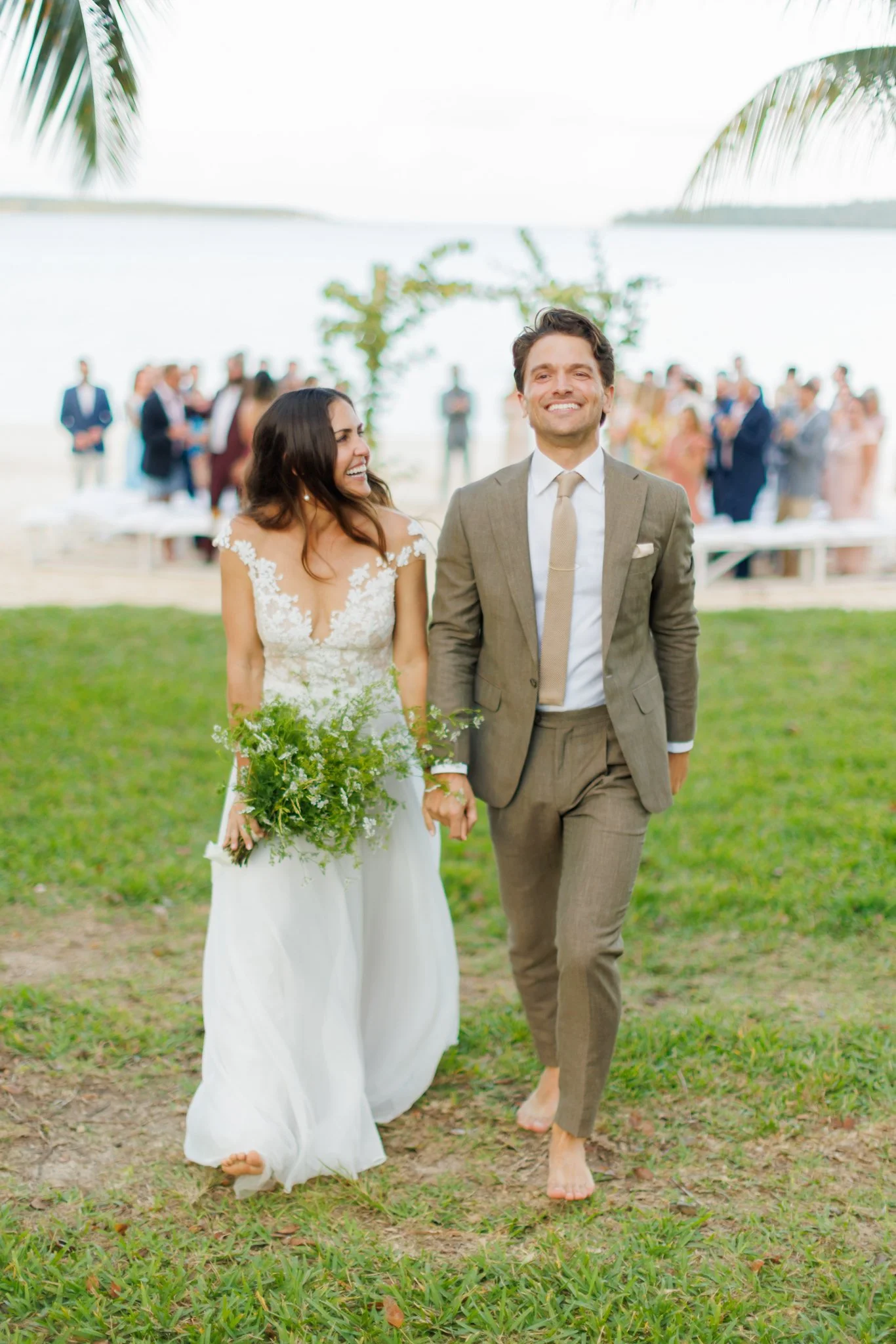 A newly married couple walking hand in hand barefoot on the grass at their outdoor wedding ceremony by a lake, with guests in the background at The Other Side Hotel Eleuthera during a luxury Bahamas destination wedding.