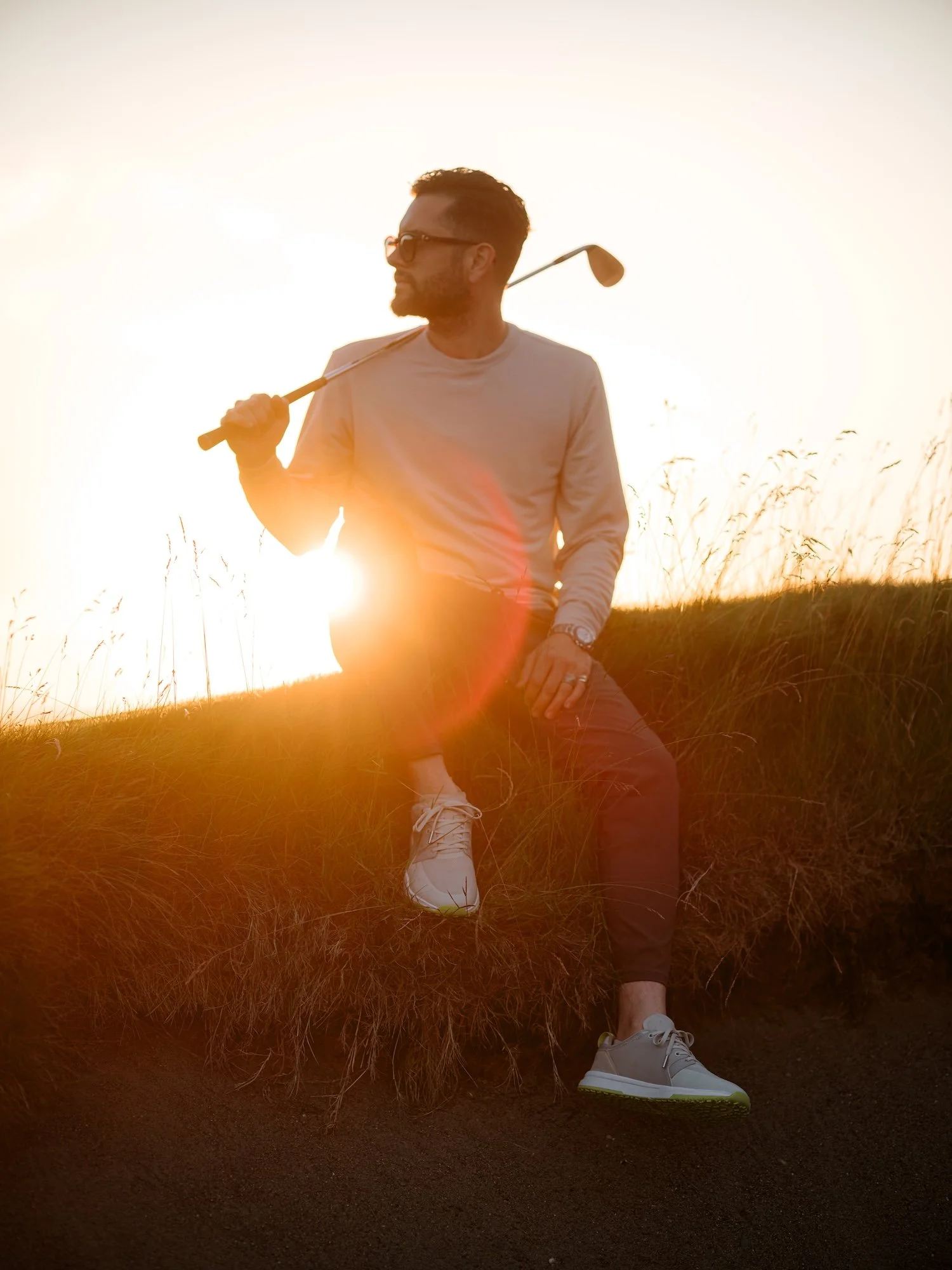 Man wearing sunglasses holding a golf club walking on a grassy hill at sunset.