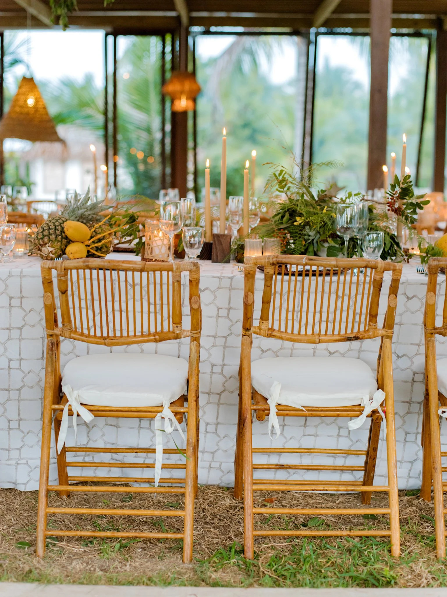 Decorative table setting at an outdoor event with bamboo chairs, white cushions, tropical fruits, greenery, candles, and glassware.