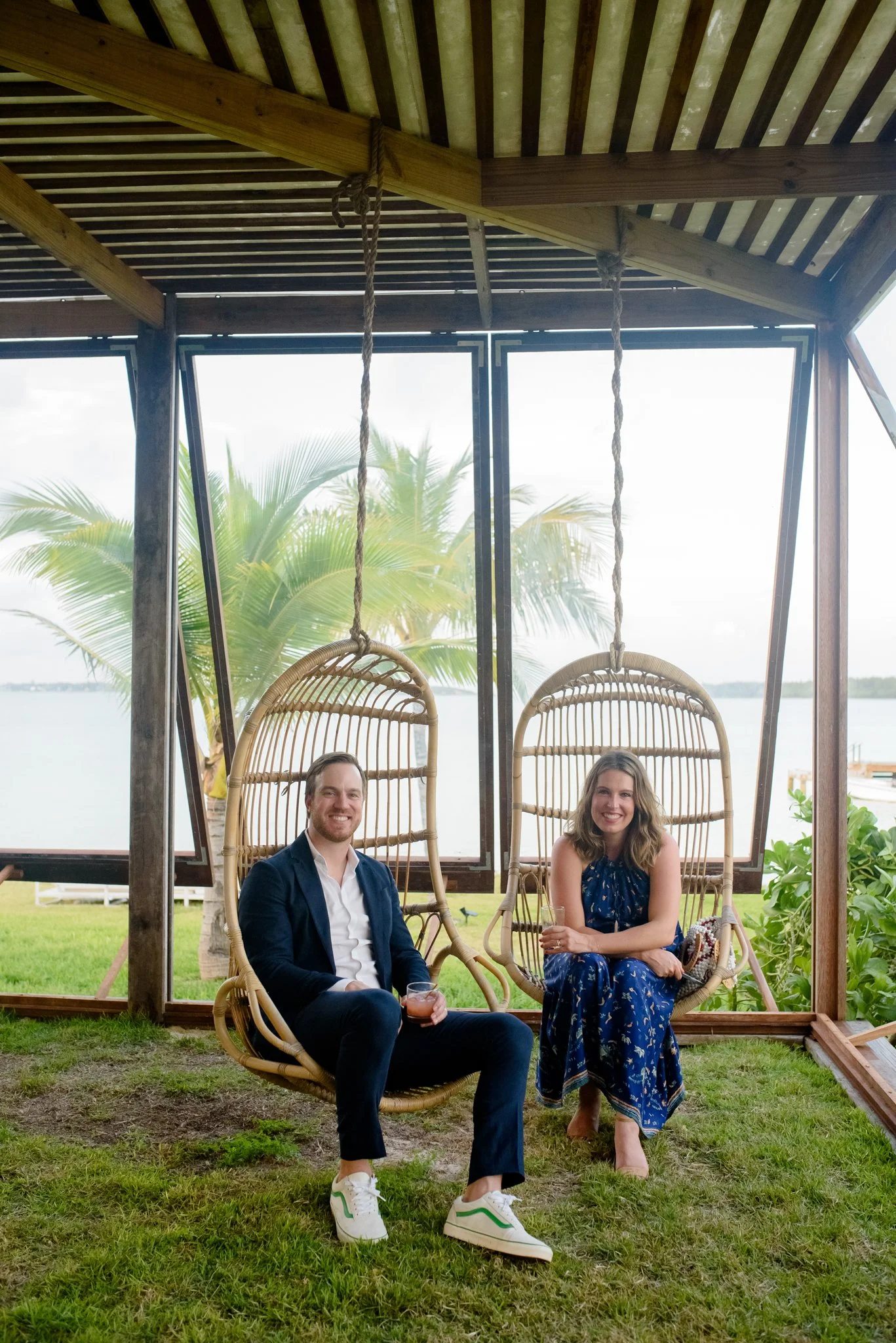A man and a woman sitting on hanging rattan chairs in a screened outdoor area, with a tropical view of palm trees, water, and cloudy sky in the background.