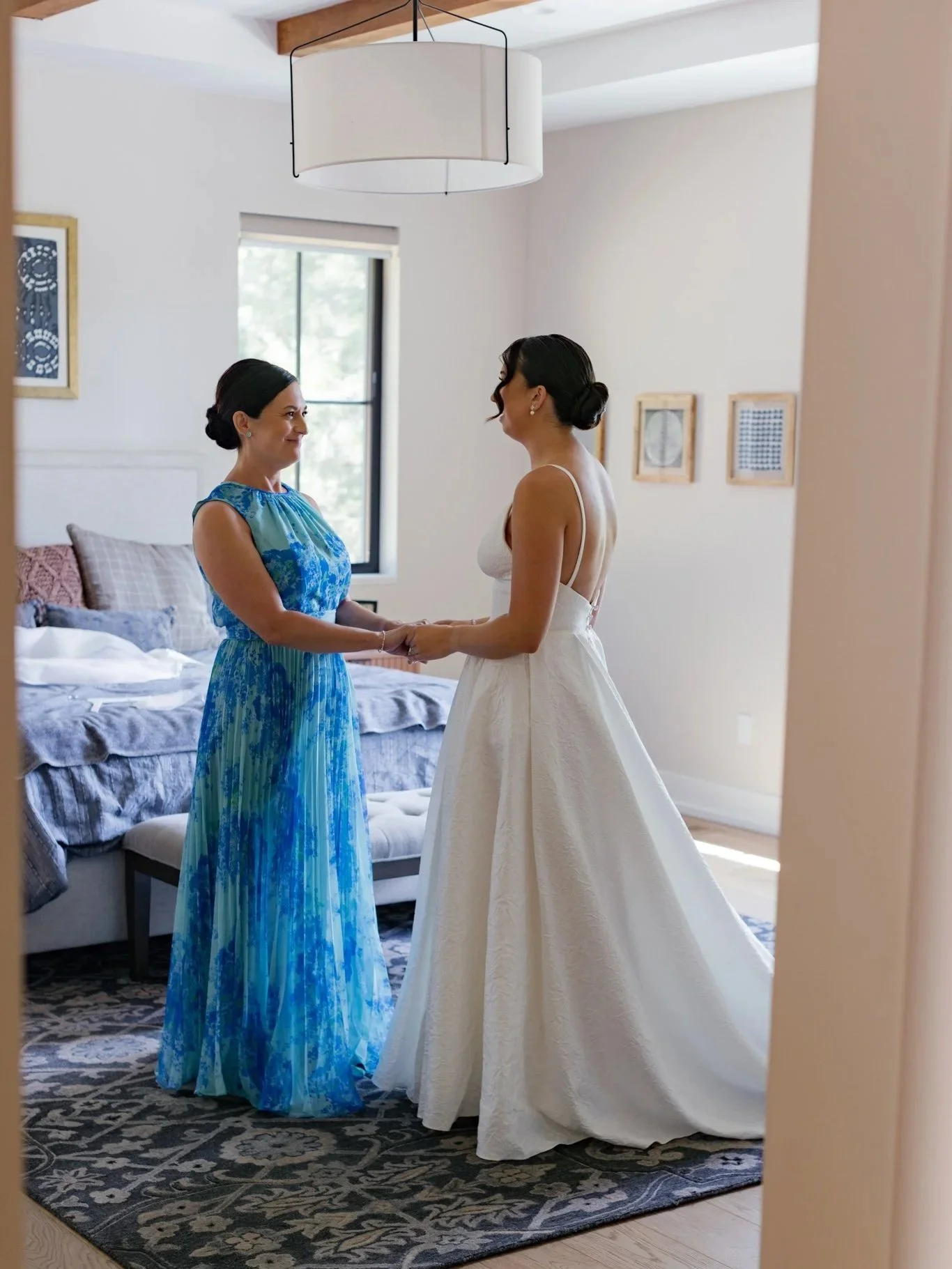 Two women, one in a white wedding dress and the other in a blue patterned dress, are holding hands and smiling at each other in a well-lit bedroom.