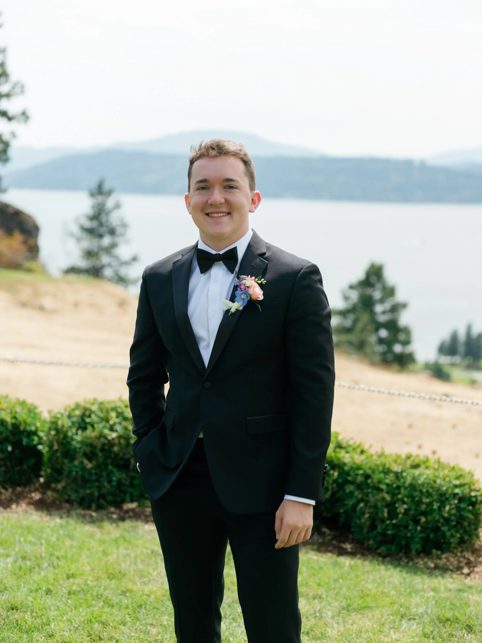 A young man in a black tuxedo with a bowtie, standing outdoors near water with mountains and trees in the background, smiling at the camera at Elk Point summer wedding above Lake Coeur d’Alene.