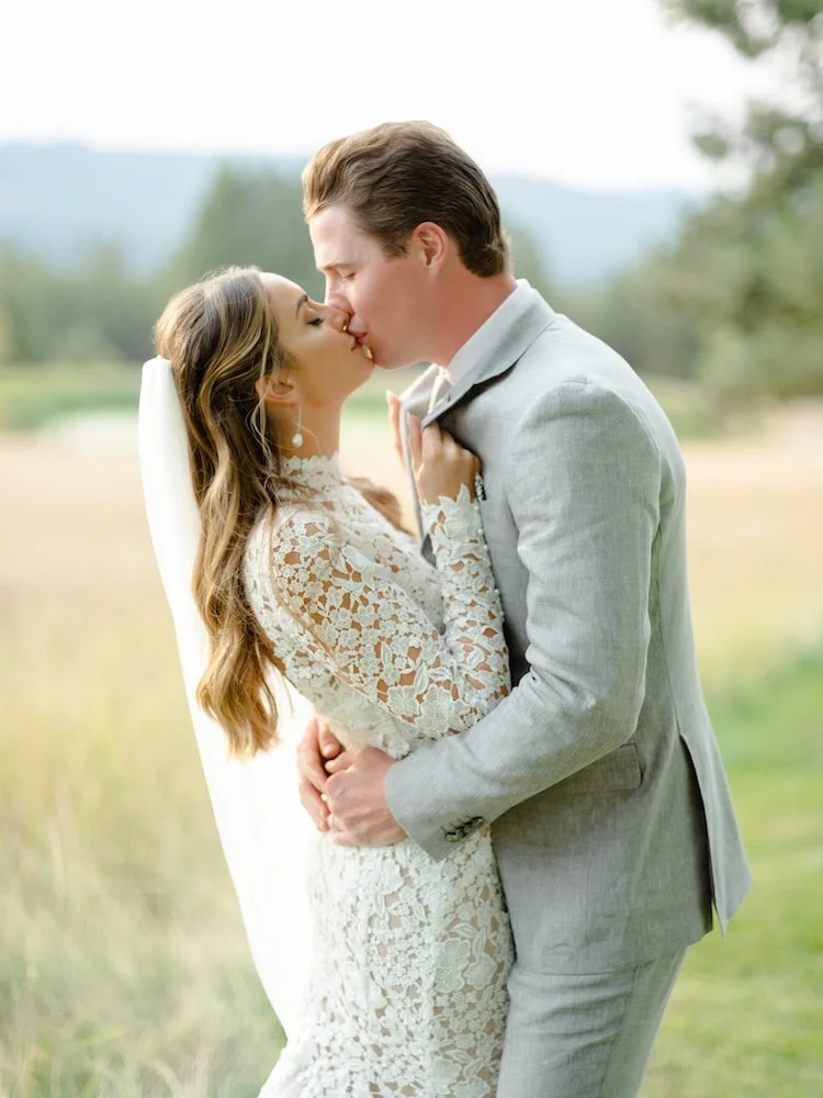 A bride and groom sharing a kiss outdoors during their wedding, with the bride wearing a lace dress and veil, and the groom in a light gray suit.