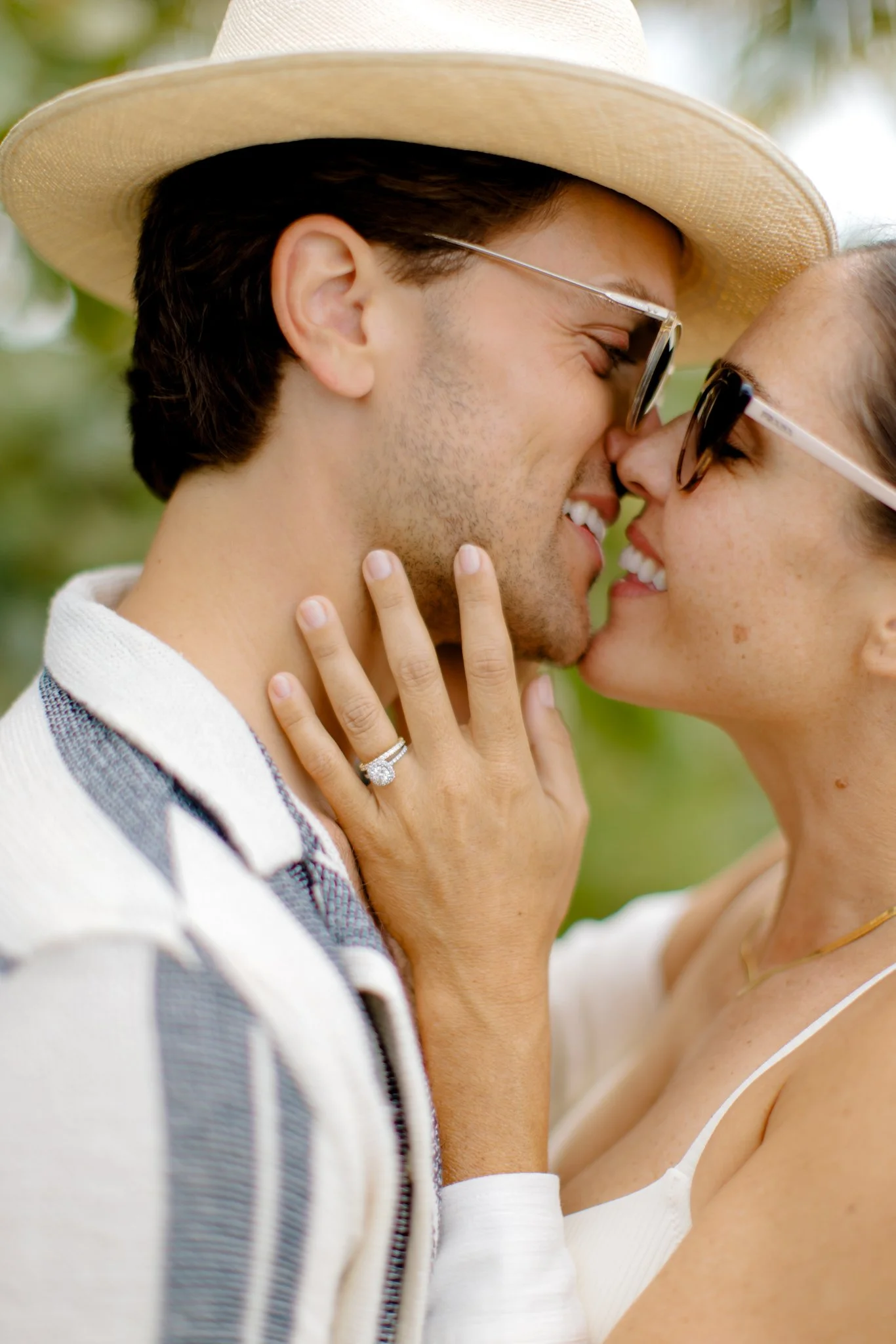 A couple is sharing a close, intimate moment outdoors, touching noses and smiling, with the woman gently holding the man's face. The woman is wearing sunglasses, a white top, and a ring, while the man is wearing a straw hat, glasses, and a striped jacket.