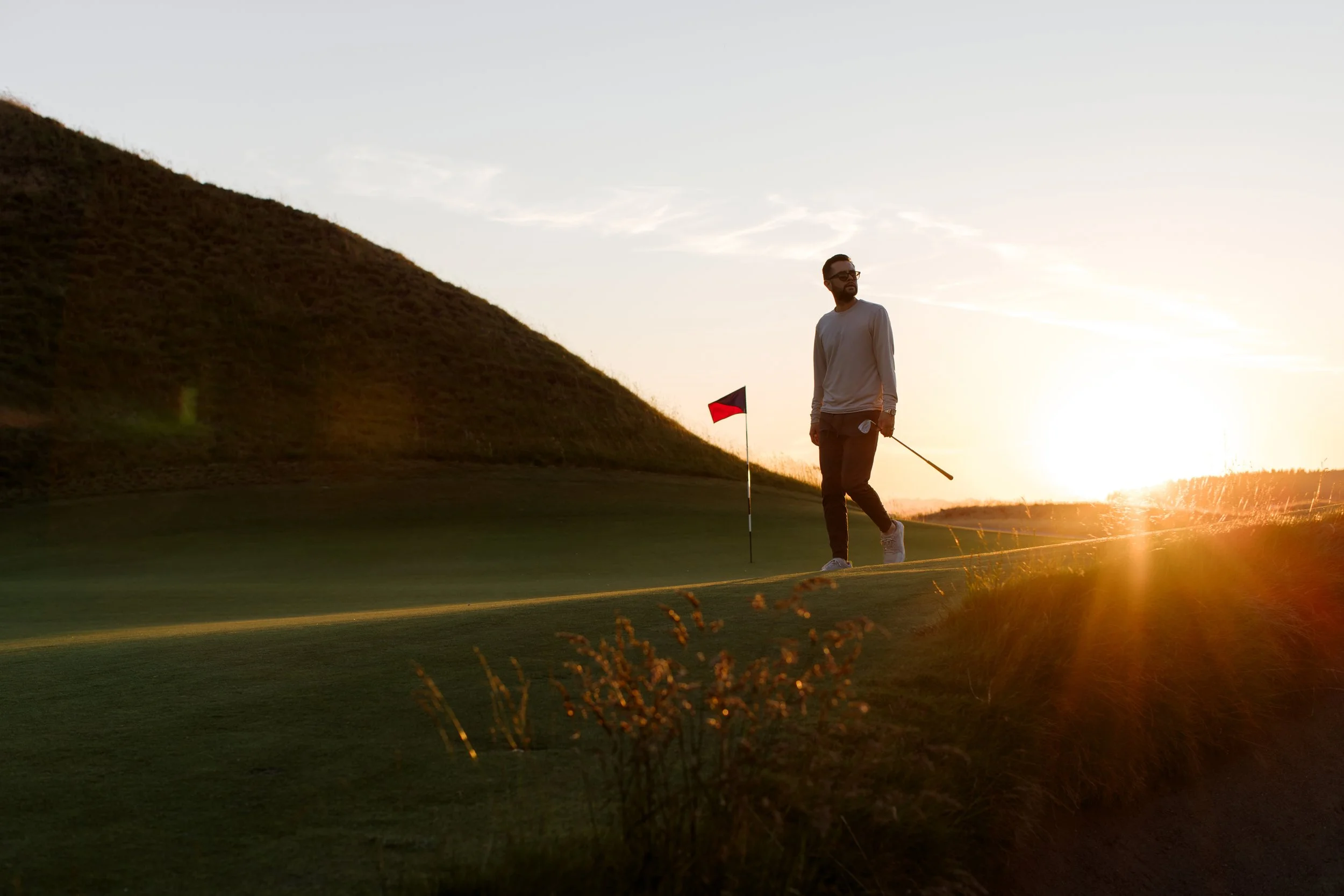 A man standing on a golf course at sunset, holding a golf club, with a flagstick nearby, hills in the background, and the sun setting in the distance.