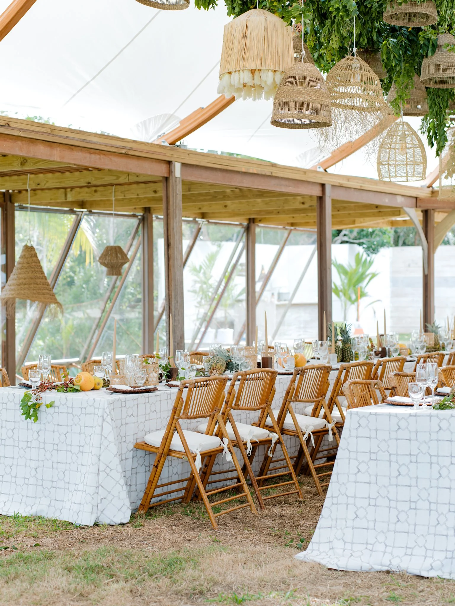 Decorated outdoor dining area with wooden chairs, white tablecloths, and tropical centerpieces under hanging lamps.