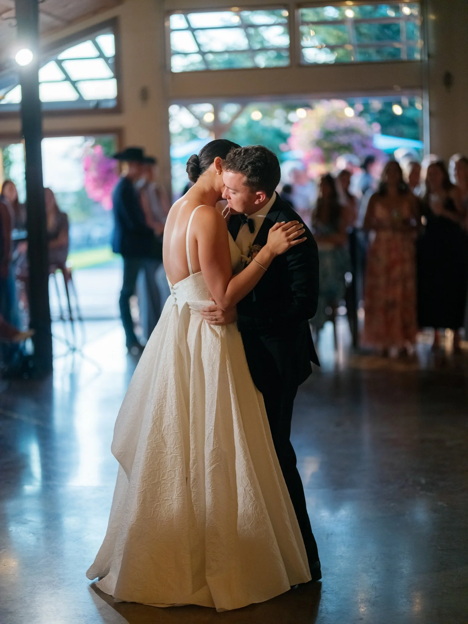 A bride and groom sharing a dance during their wedding reception inside a venue with large windows and hanging decorations, with guests watching in the background at Elk Point wedding in Coeur d’Alene.