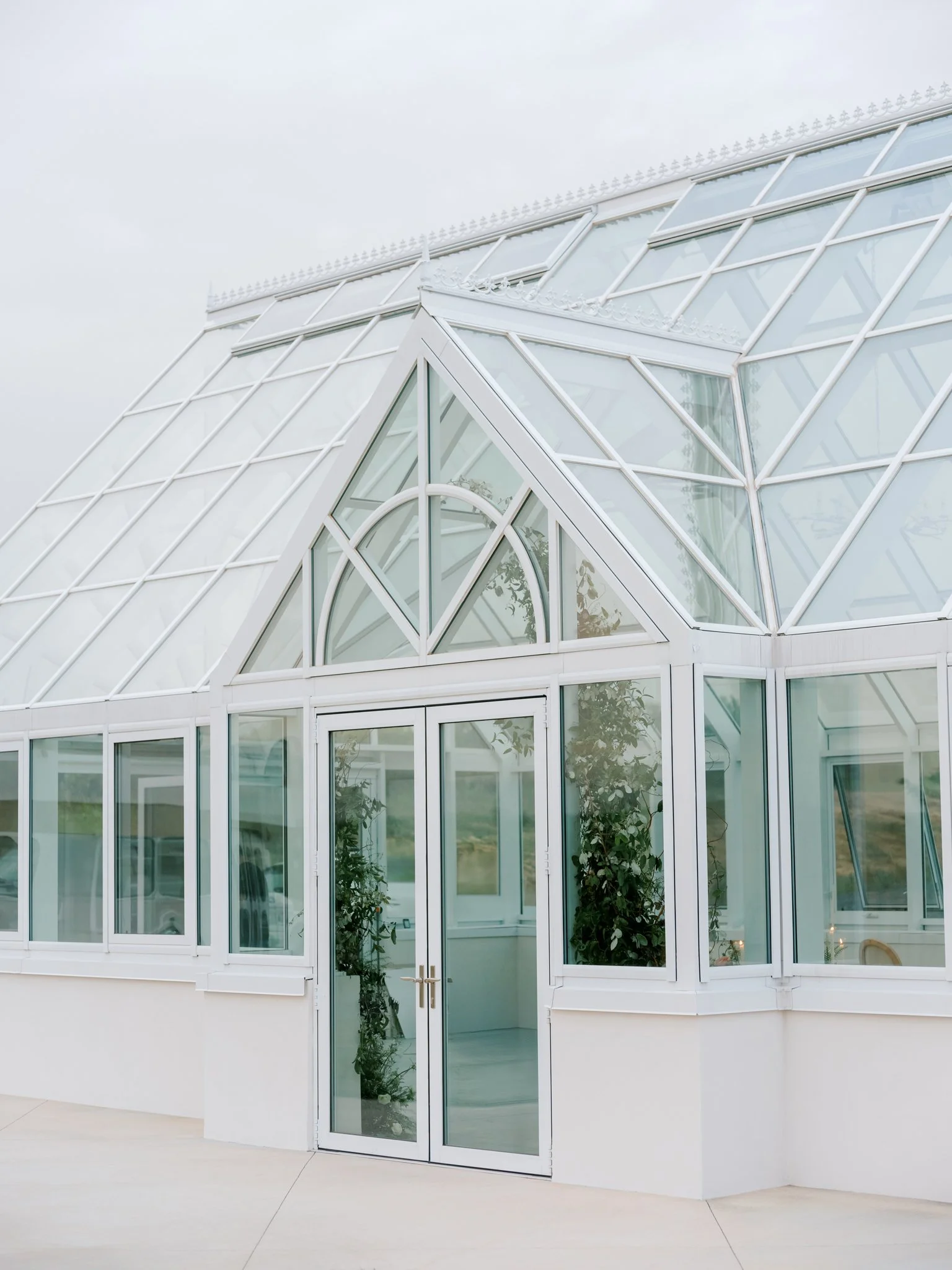 A glass greenhouse with white framing and a double door entrance, featuring triangular and rectangular glass panels, with plants visible inside at Greenhouse on Harvard near Spokane, Washington