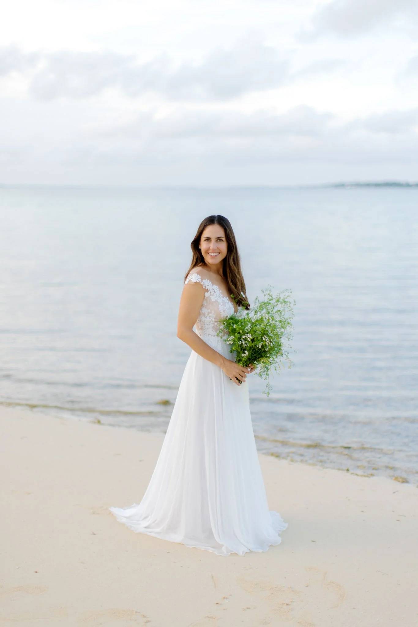 A woman in a white wedding dress holding a bouquet of flowers on a sandy beach near the water.