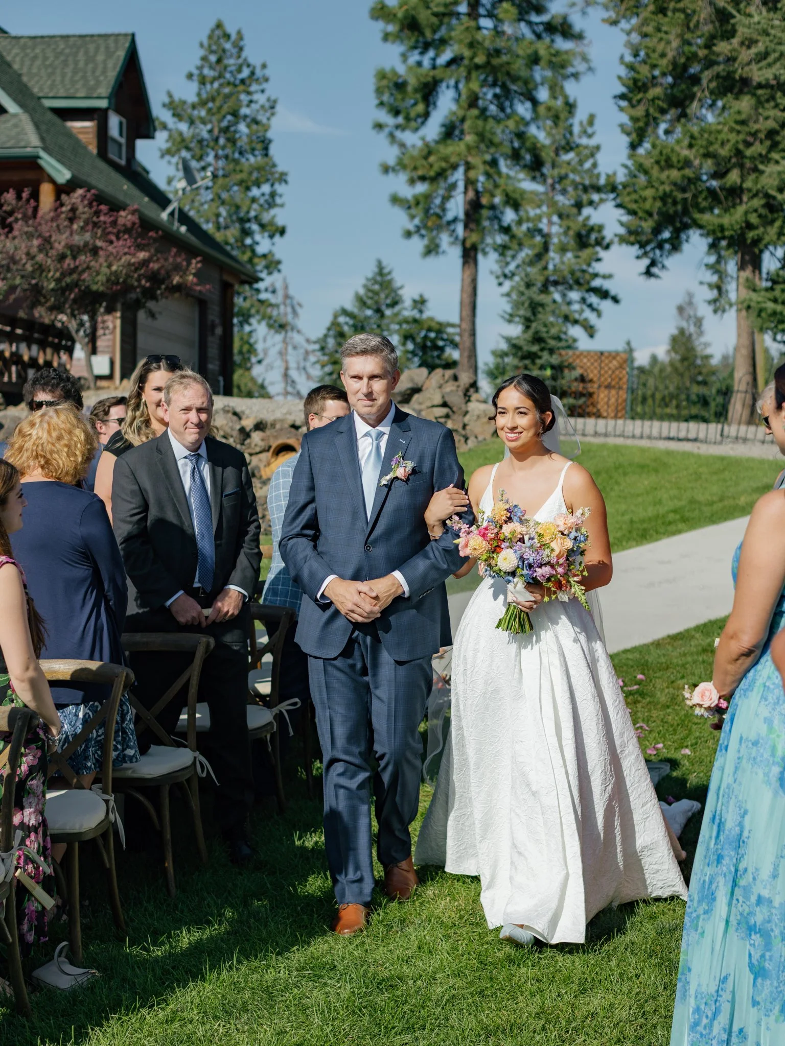 A bride walking down an outdoor aisle, accompanied by a man, possibly her father, during a wedding ceremony. The bride is holding a colorful bouquet, and there are guests standing and sitting on chairs on either side of the aisle. The scene is set in a lush, green area with tall trees and a house in the background.