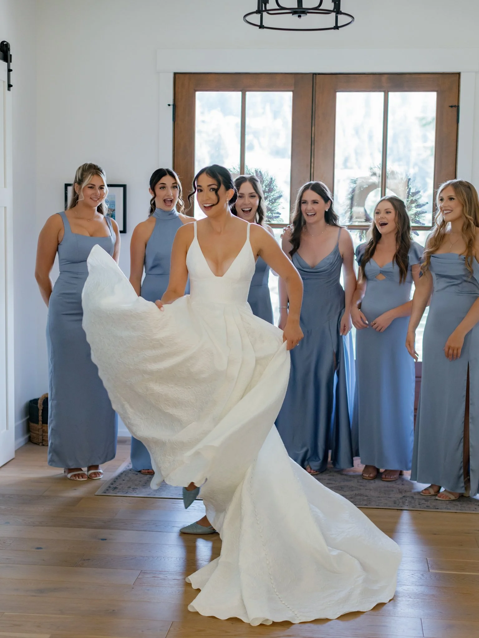 A bride in a white wedding dress posing happily in front of her bridesmaids, who are dressed in matching light blue dresses, inside a well-lit room with large windows at Elk Point wedding above Lake Coeur d’Alene.