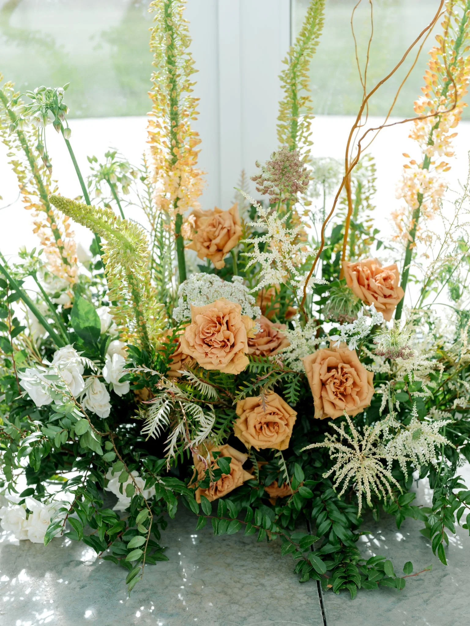 A floral arrangement of peach roses, white flowers, and greenery with tall, light-colored flowers, set against a bright window at Greenhouse on Harvard near Spokane, Washington.