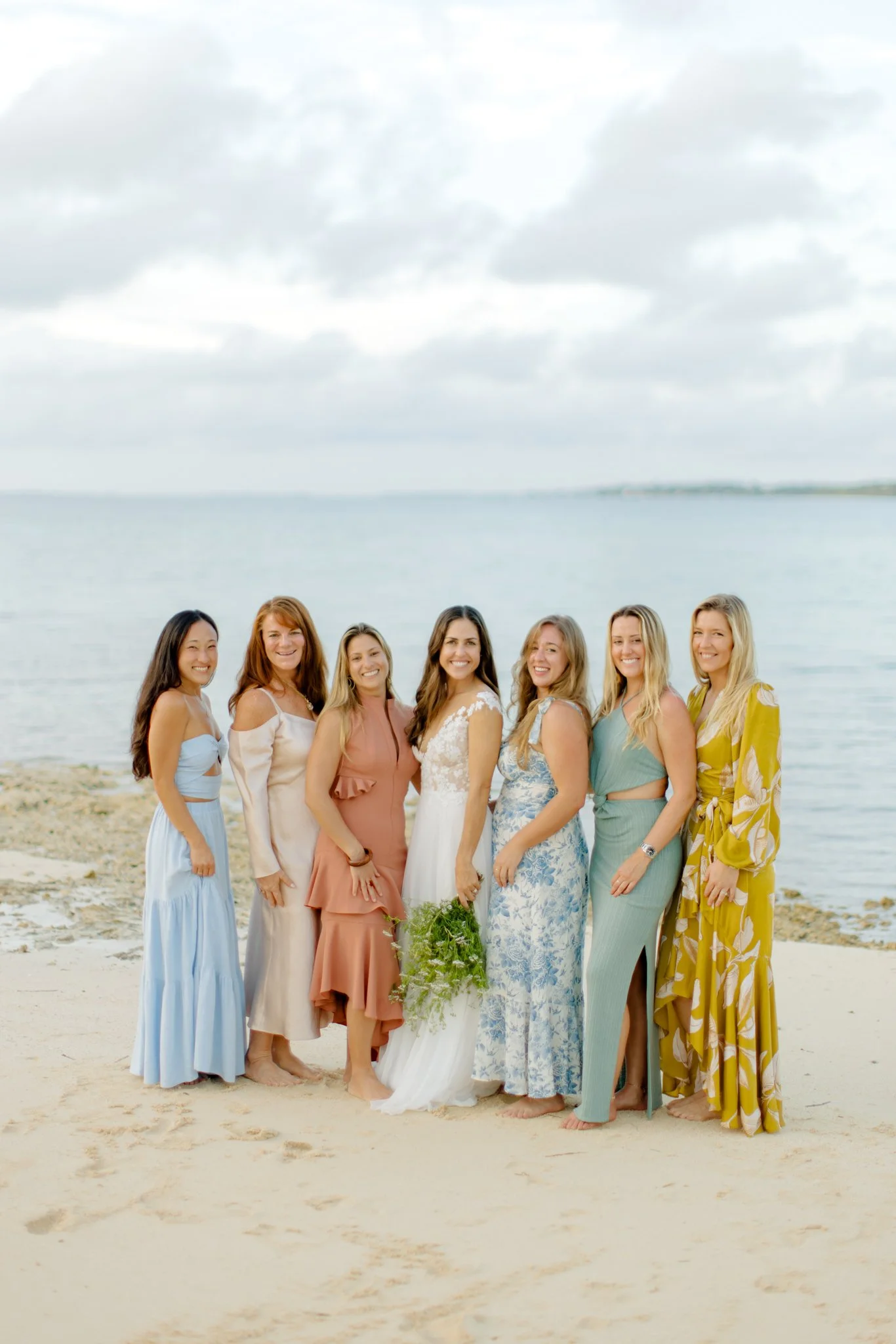 A group of seven women in colorful dresses standing on a beach with water and cloudy sky in the background, some women holding greenery, celebrating or attending a special occasion.