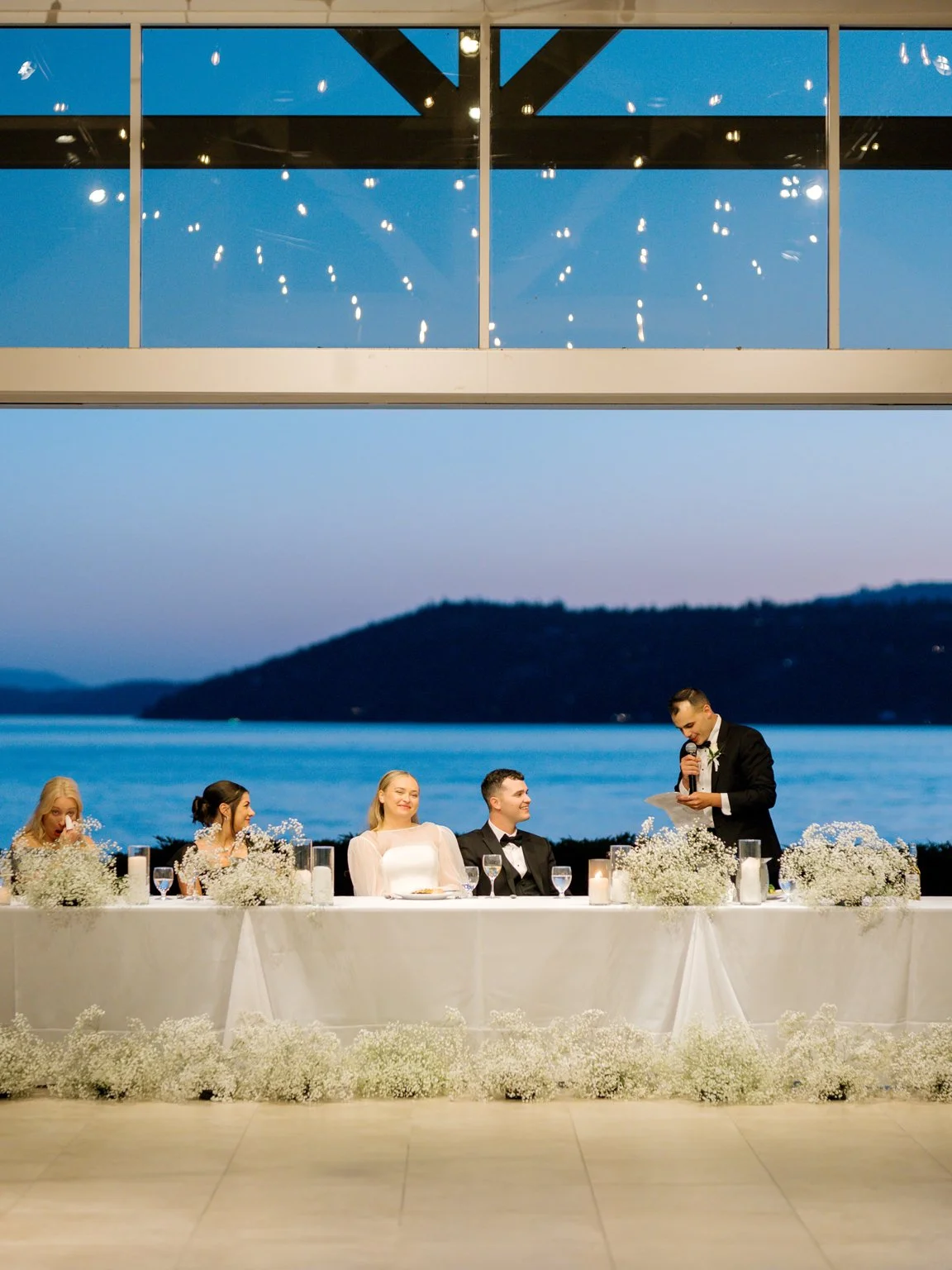 Wedding reception with bride and groom seated at a decorated table by a large window overlooking a lake at dusk, with a man giving a toast.