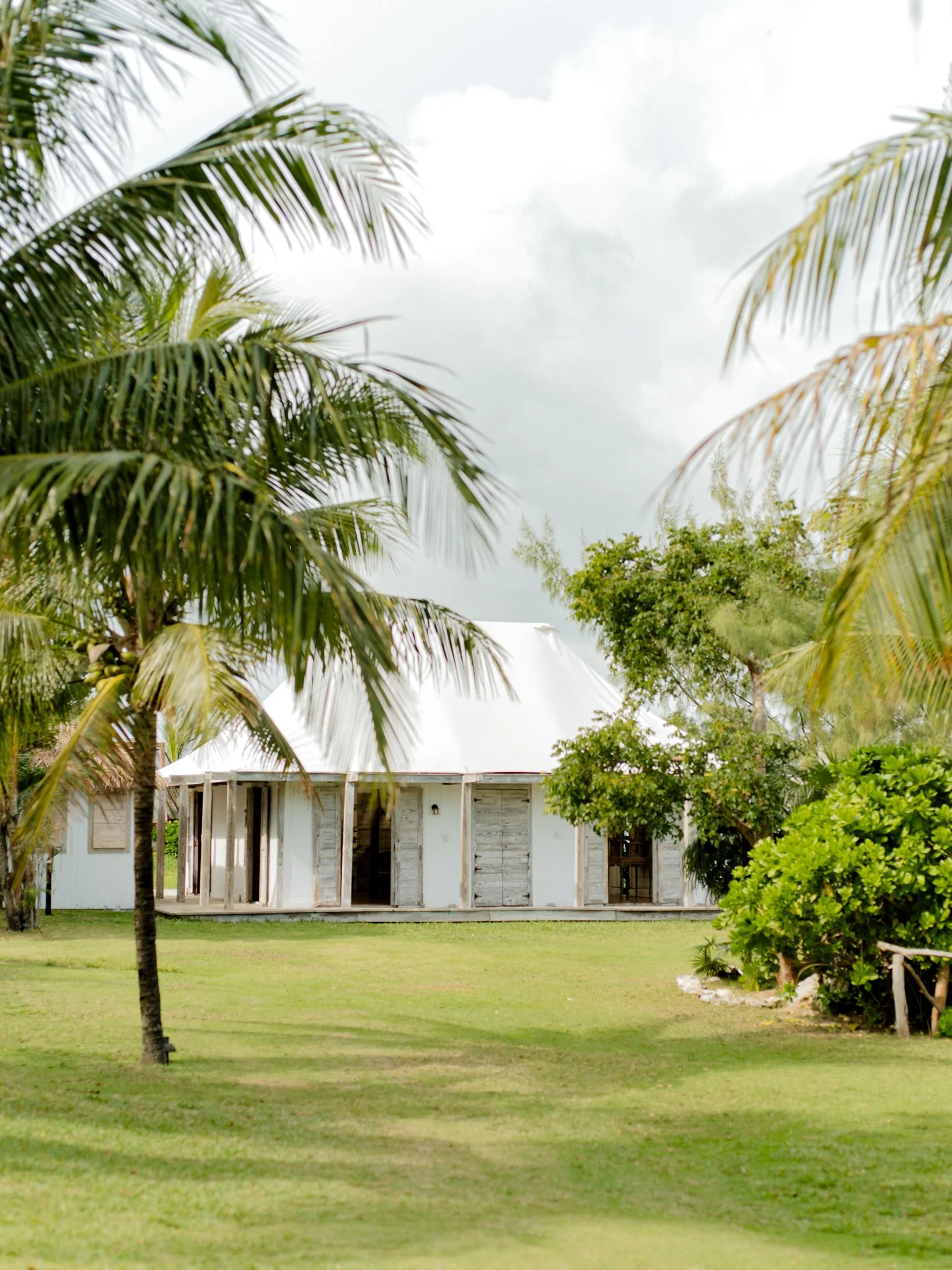 A tropical house with a white roof surrounded by palm trees and green grass, under a cloudy sky at The Other Side Hotel Eleuthera during a luxury Bahamas destination wedding.