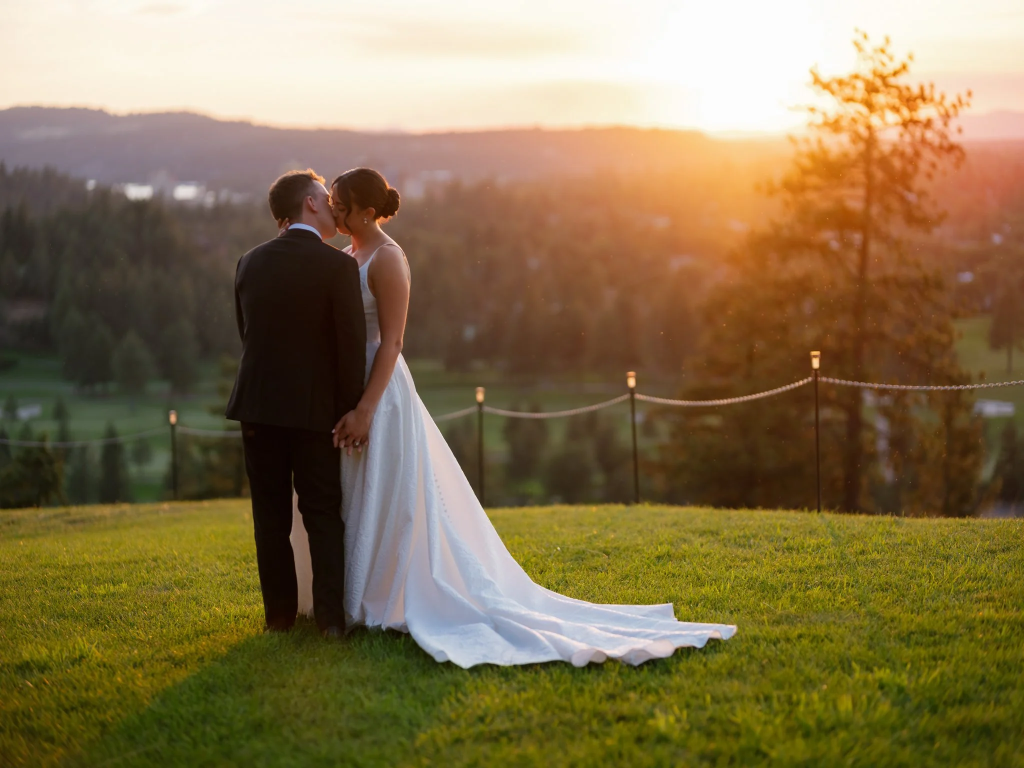 A bride and groom kissing outdoors at sunset, holding hands on a grassy area with a scenic background of trees and distant hills in Coeur d'Alene, Idaho.
