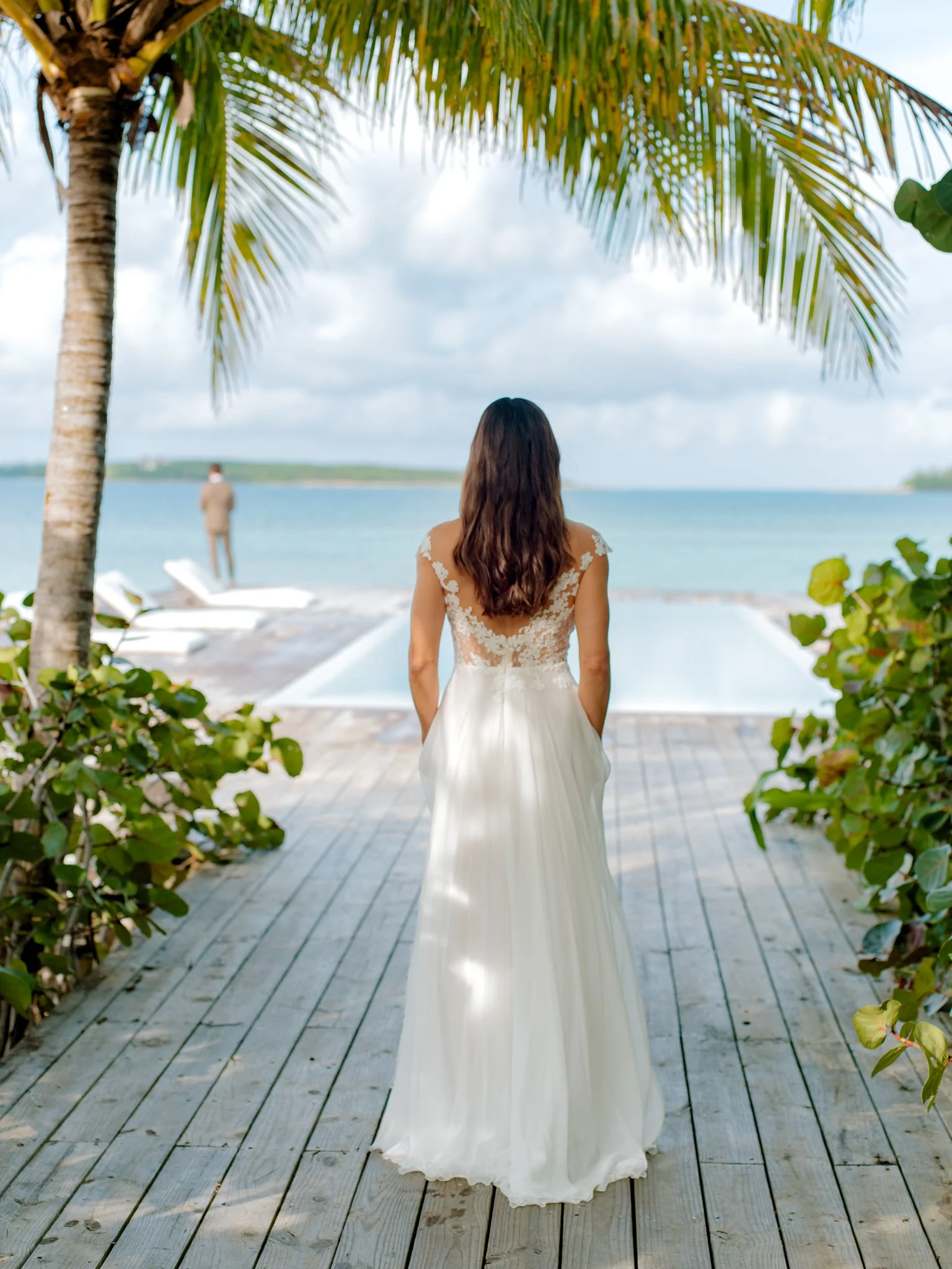 A woman in a wedding dress stands on a wooden pathway overlooking the ocean, with palm trees and bushes framing the scene, and a person in a suit in the distance near lounge chairs by the water.