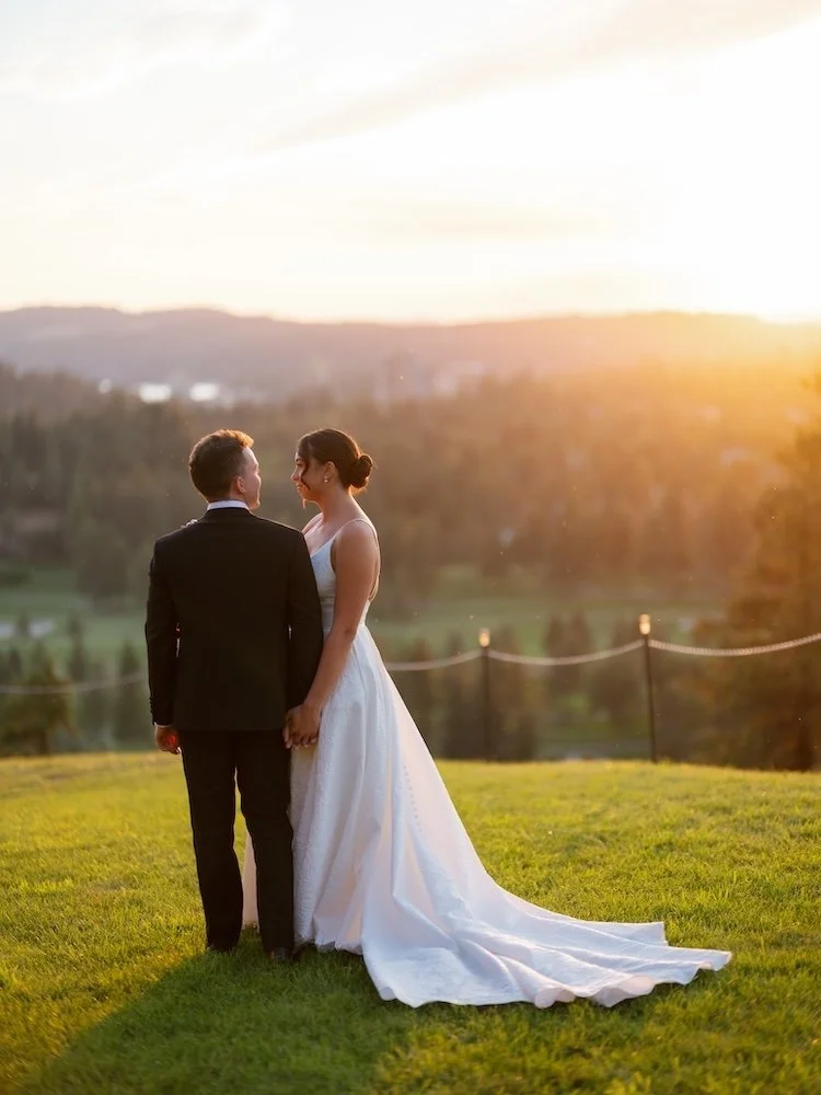 A bride and groom standing hand in hand on a lush green field at sunset, with a scenic landscape of trees and hills in the background.