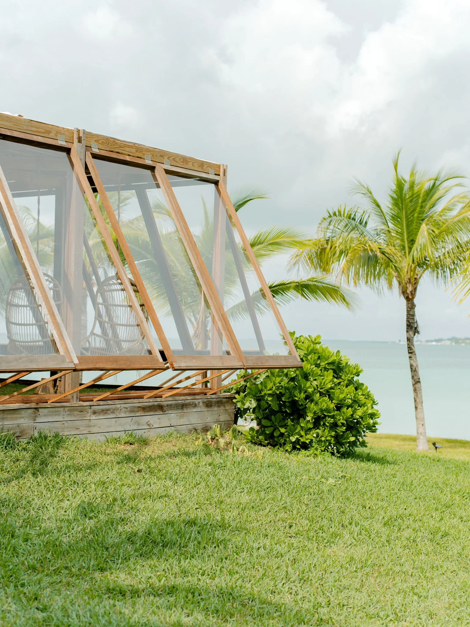 A wooden outdoor patio structure with glass walls, tropical plants including palm trees, and a grassy area overlooking water with cloudy sky.