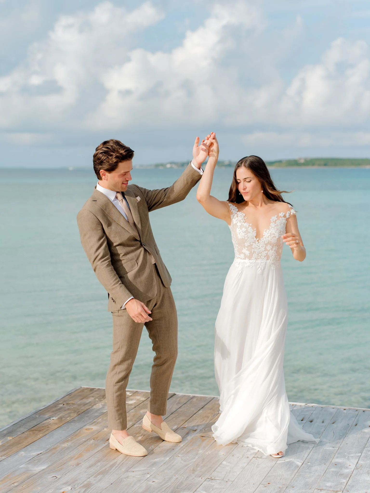 A couple dancing on a wooden pier by the water, with the man wearing a brown suit and the woman in a white wedding dress at The Other Side Hotel Eleuthera during a luxury Bahamas destination wedding.