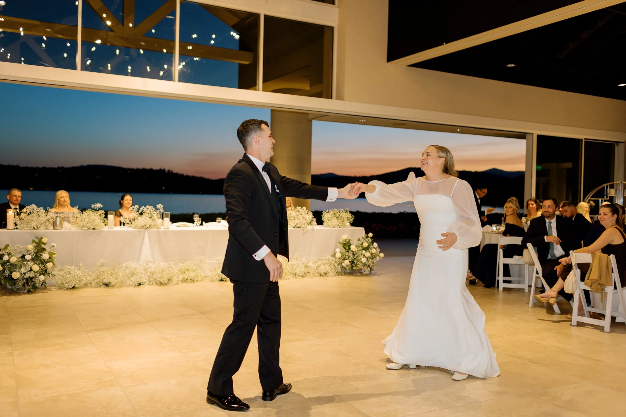 A bride and groom dancing at their wedding reception with guests seated at tables in the background and a scenic sunset view outside at the Hagadone Event Center in Coeur d'Alene, Idaho.